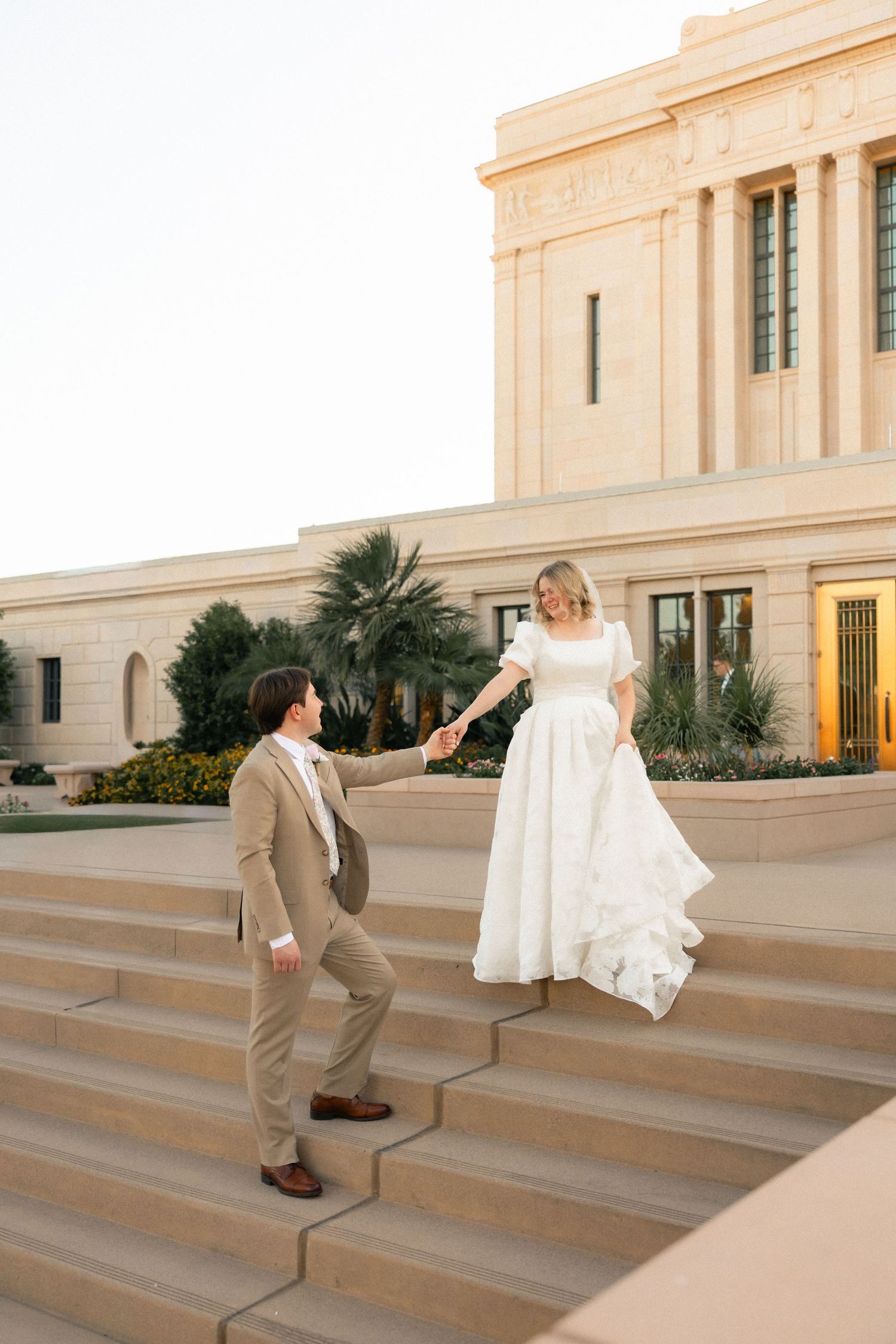 Bride and groom hold hands on temple steps, she in white gown, he in tan suit, elegant building backdrop.