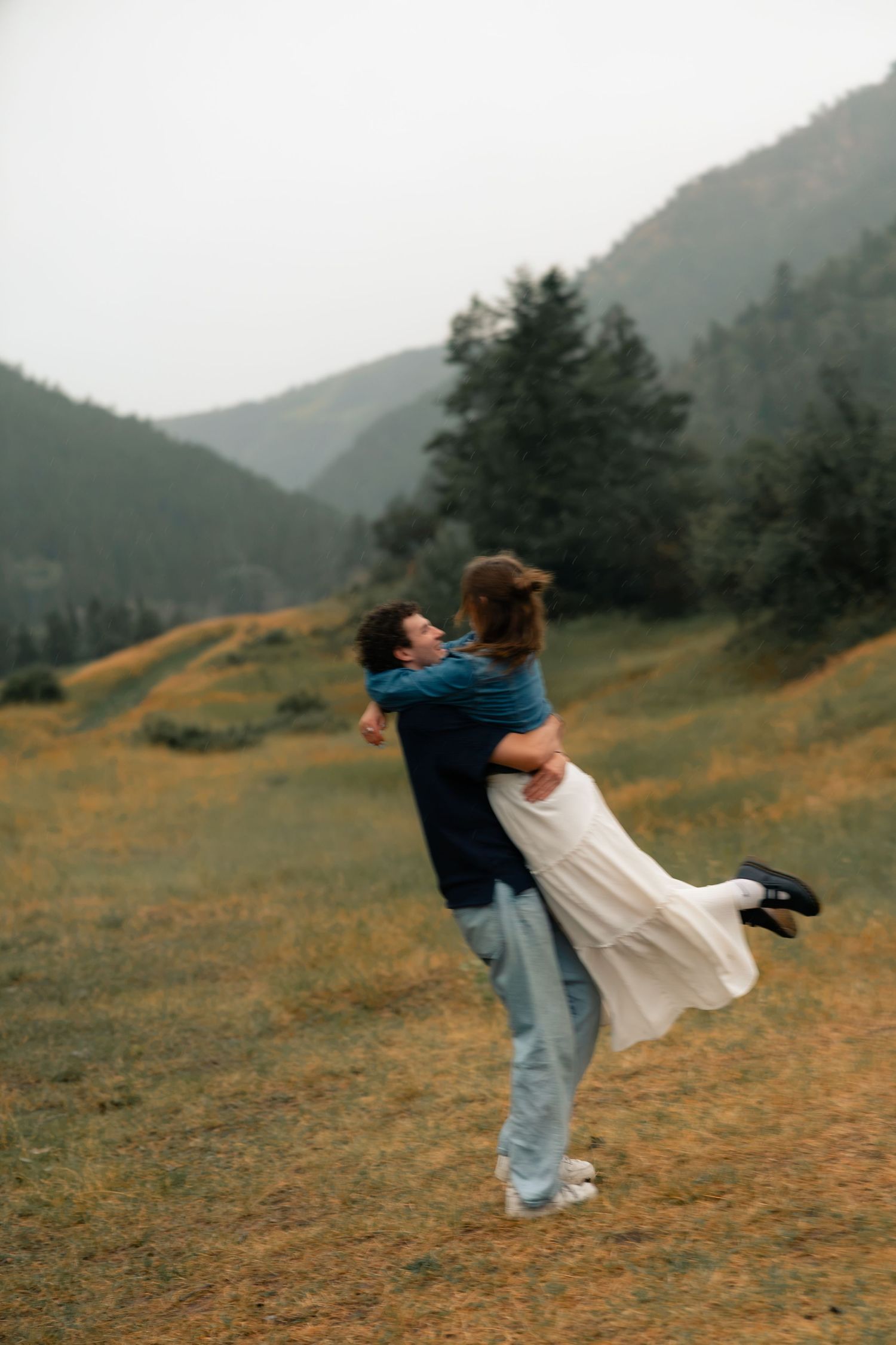 A man lifts a woman in a white dress in a mountain meadow, sharing a romantic moment amid misty green hills.