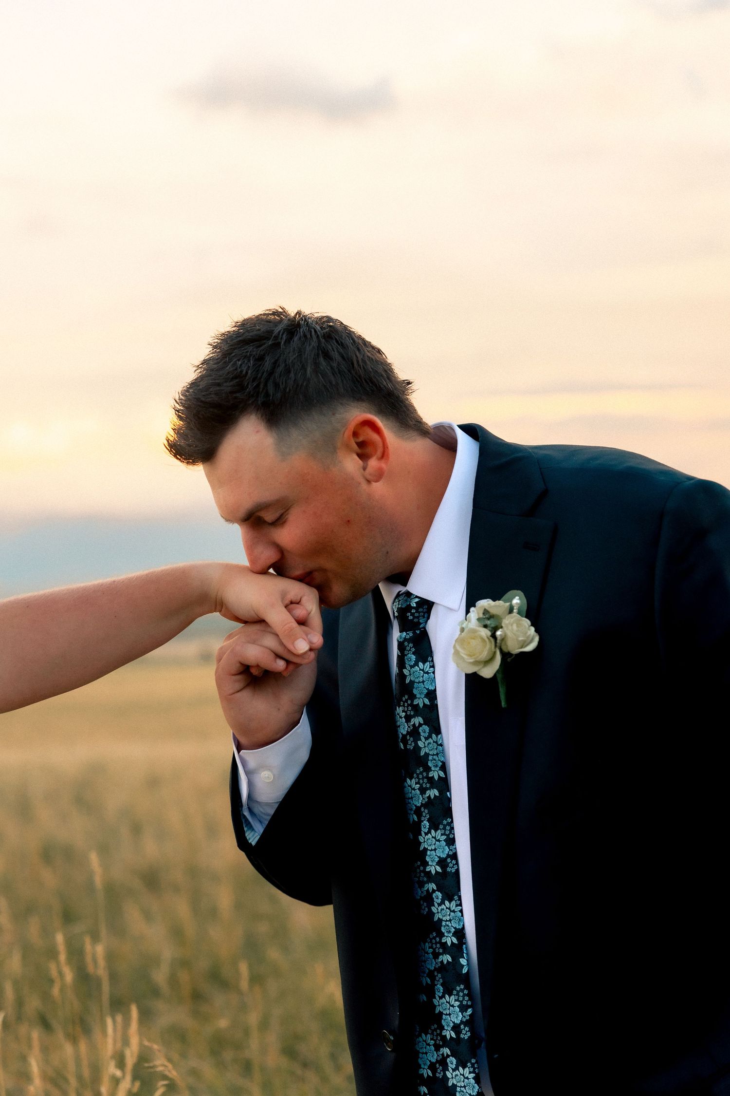 Groom in dark suit kisses bride's hand at sunset in a golden field during wedding portraits.