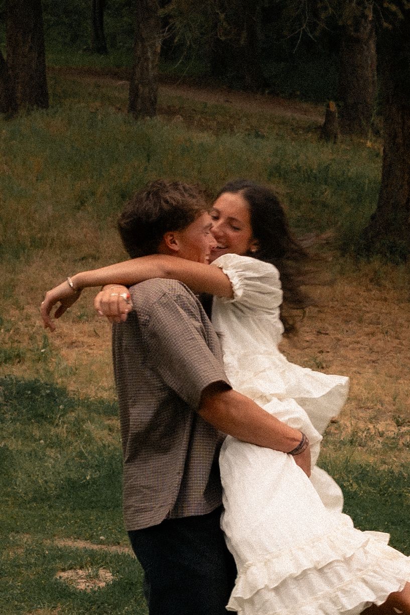 A couple embraces and dances joyfully outdoors in a forest clearing, woman in white dress, man in plaid shirt.