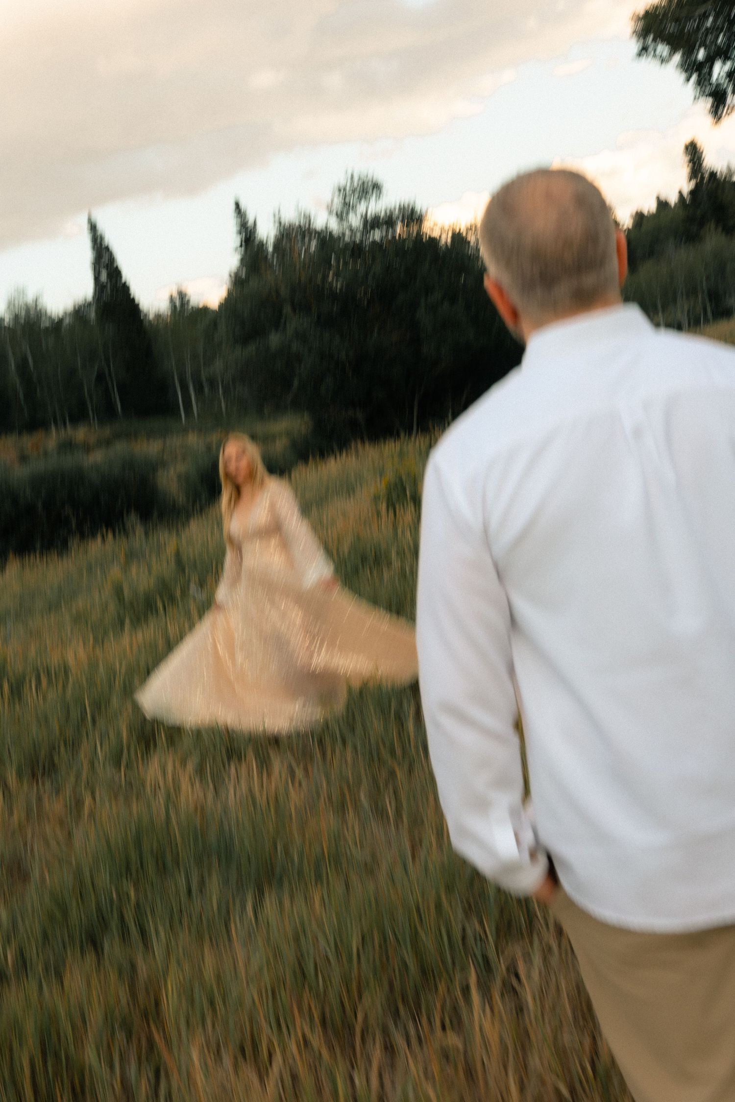 A man in a white shirt watches a woman in a flowing dress twirling in a lush green field at sunset.
