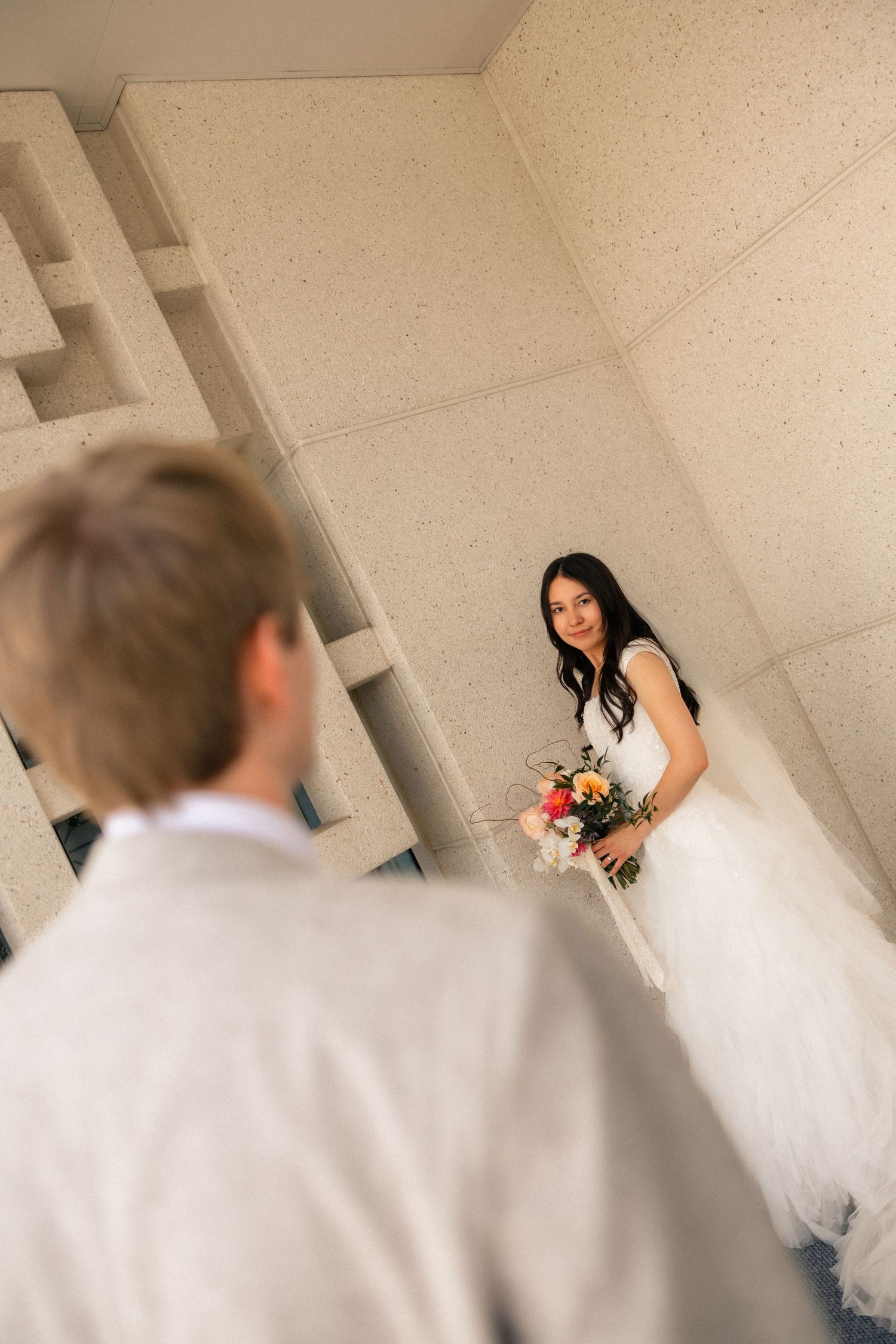 Bride in white gown holding colorful bouquet smiles at groom during wedding first look at modern building.