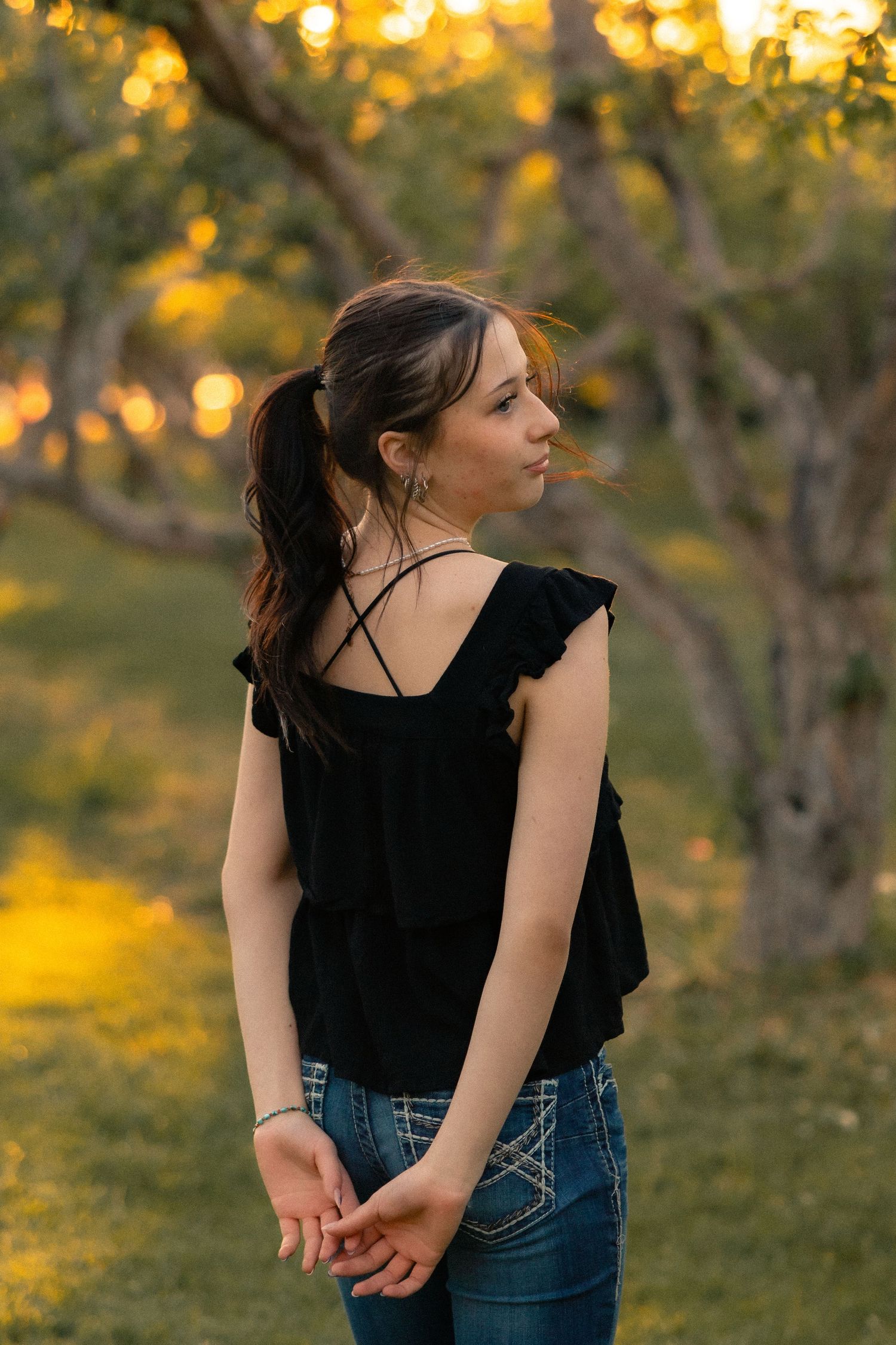 Young woman in black ruffled top and jeans stands in golden-lit orchard at sunset, hair in ponytail.