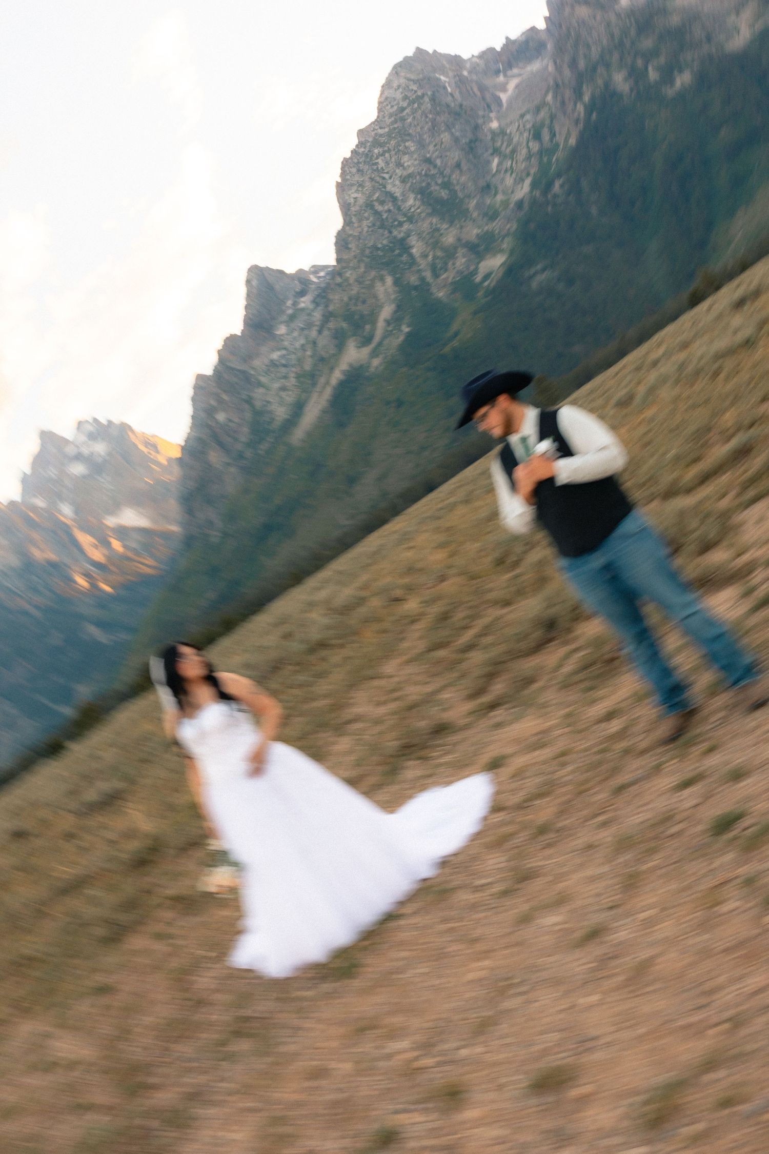 Bride in white gown and groom in cowboy hat dance on a mountain hillside with dramatic rocky peaks backdrop.