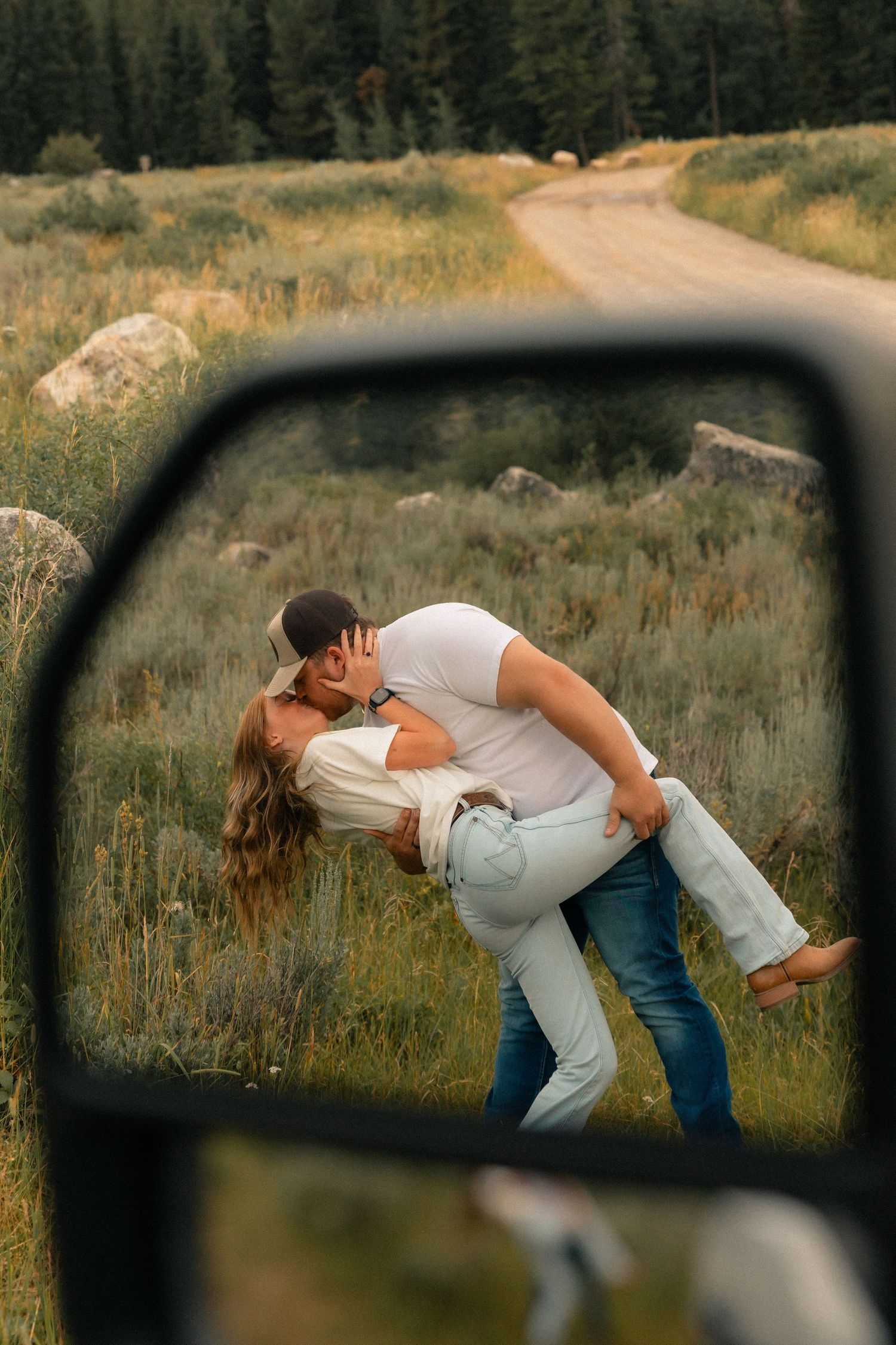 Couple sharing a romantic dip kiss in a meadow, reflected in a car mirror with a dirt road and pine trees behind them.