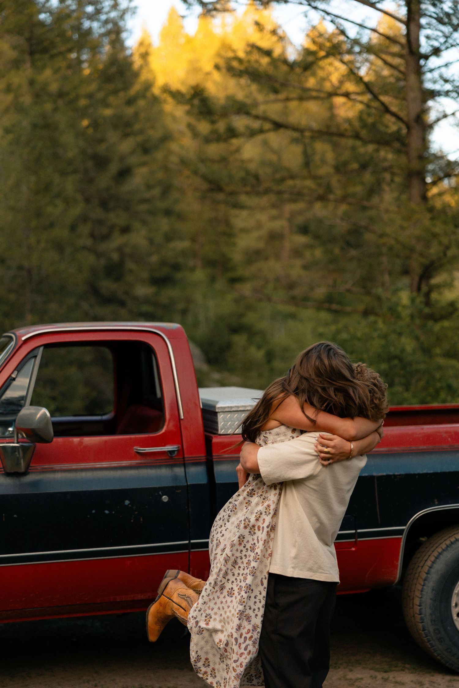 Couple embracing in a warm hug beside a classic red pickup truck with colorful autumn trees in the background.
