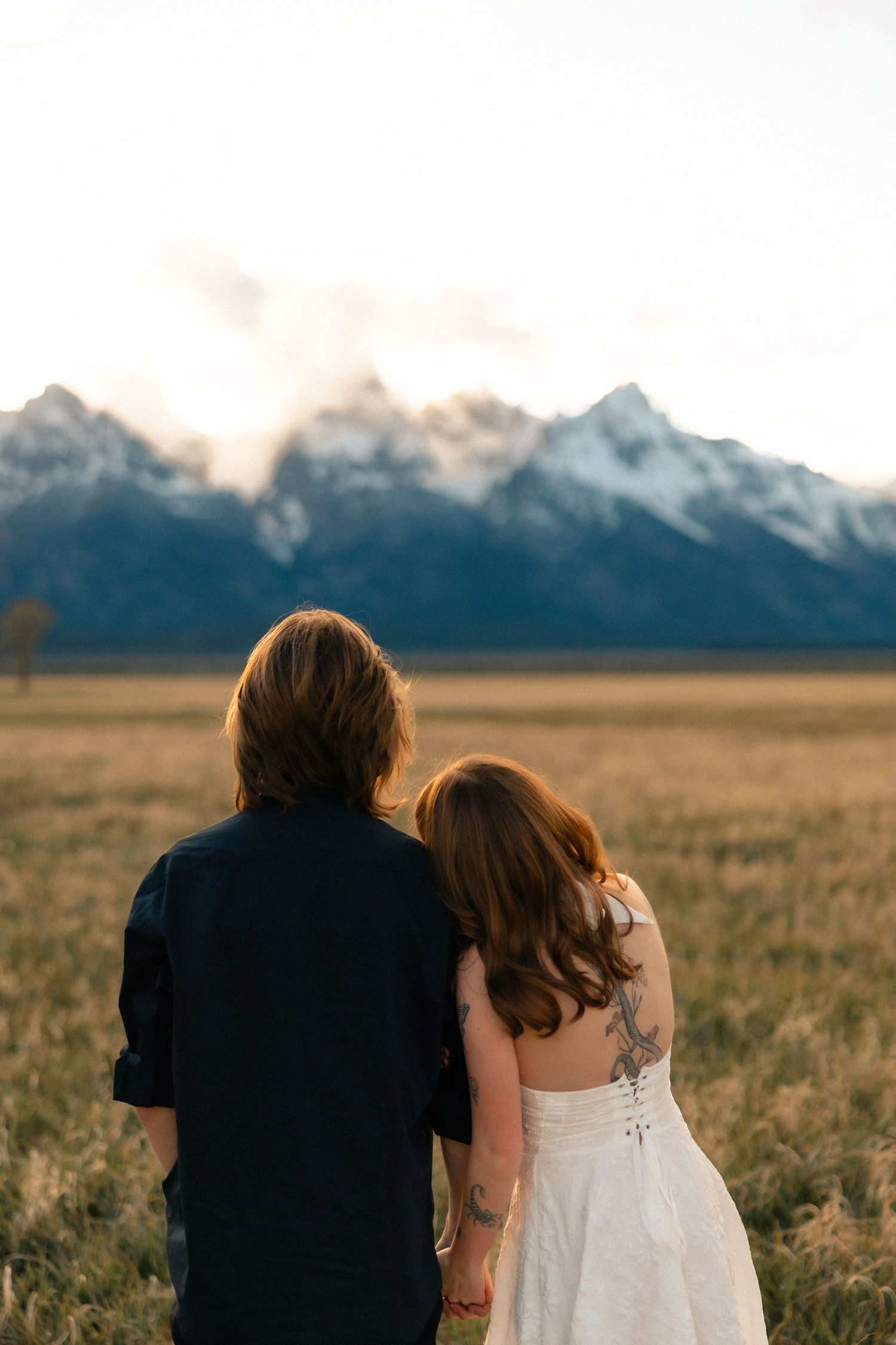 A couple holding hands gazes at snow-capped Grand Teton mountains across a golden field during their wedding day.