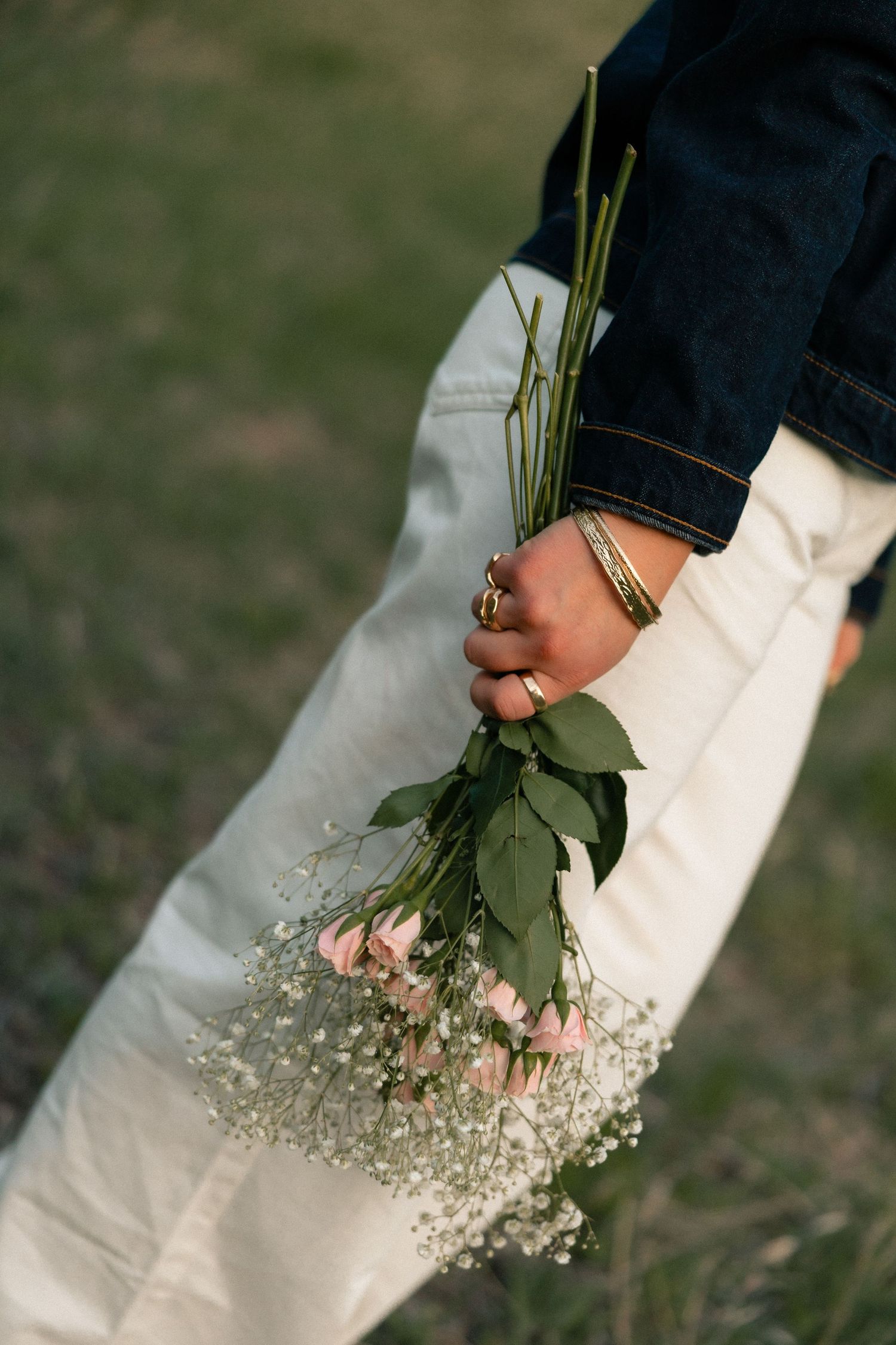 Hand holding a bouquet of pink roses and baby's breath flowers, wearing gold rings and bracelet, denim jacket.