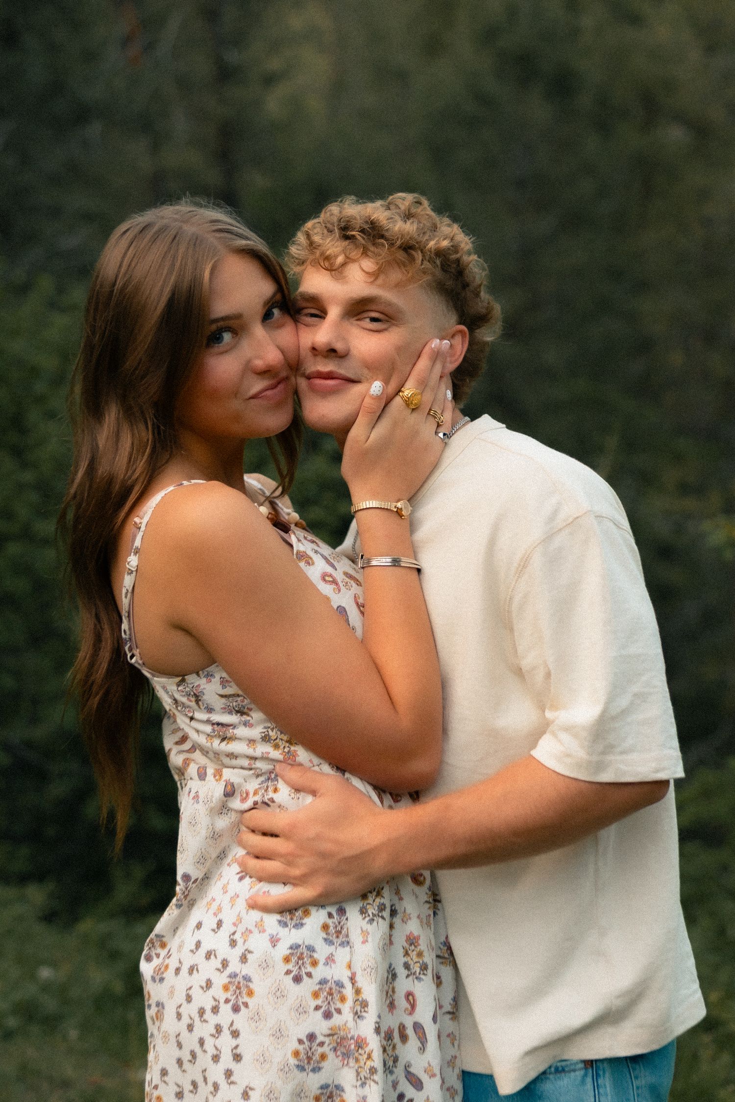 A young couple embraces outdoors, the woman in a floral dress touching the man's face, with green trees in the background.