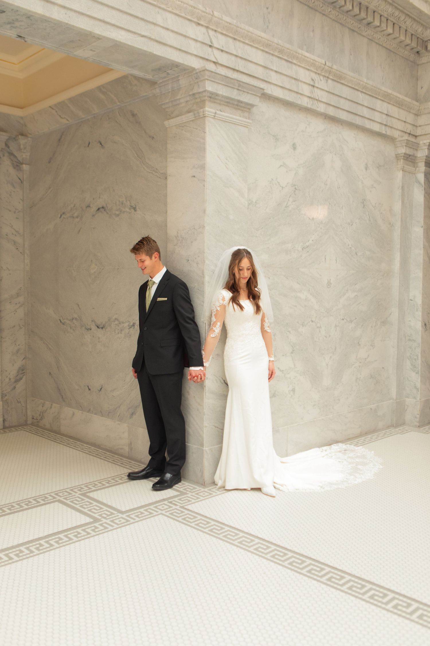 Bride in white gown and groom in black suit hold hands while standing back-to-back against marble walls.