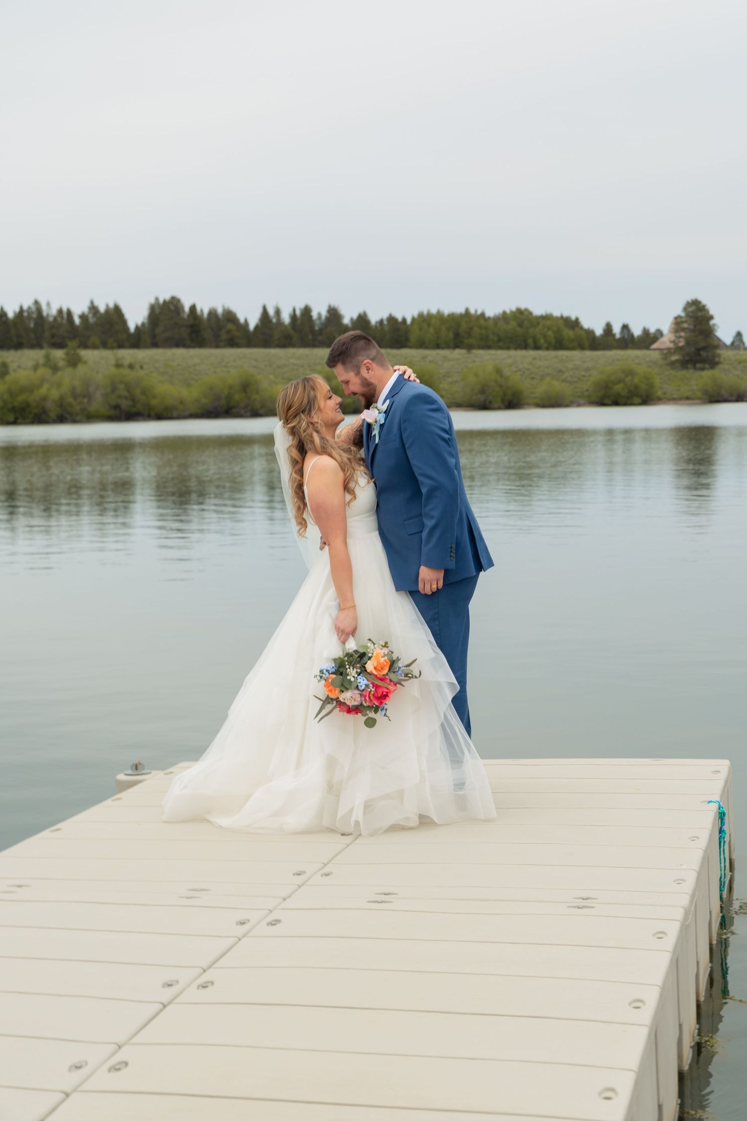 Bride and groom kissing on a dock by a serene lake, bride in white gown holding colorful bouquet, groom in blue suit.