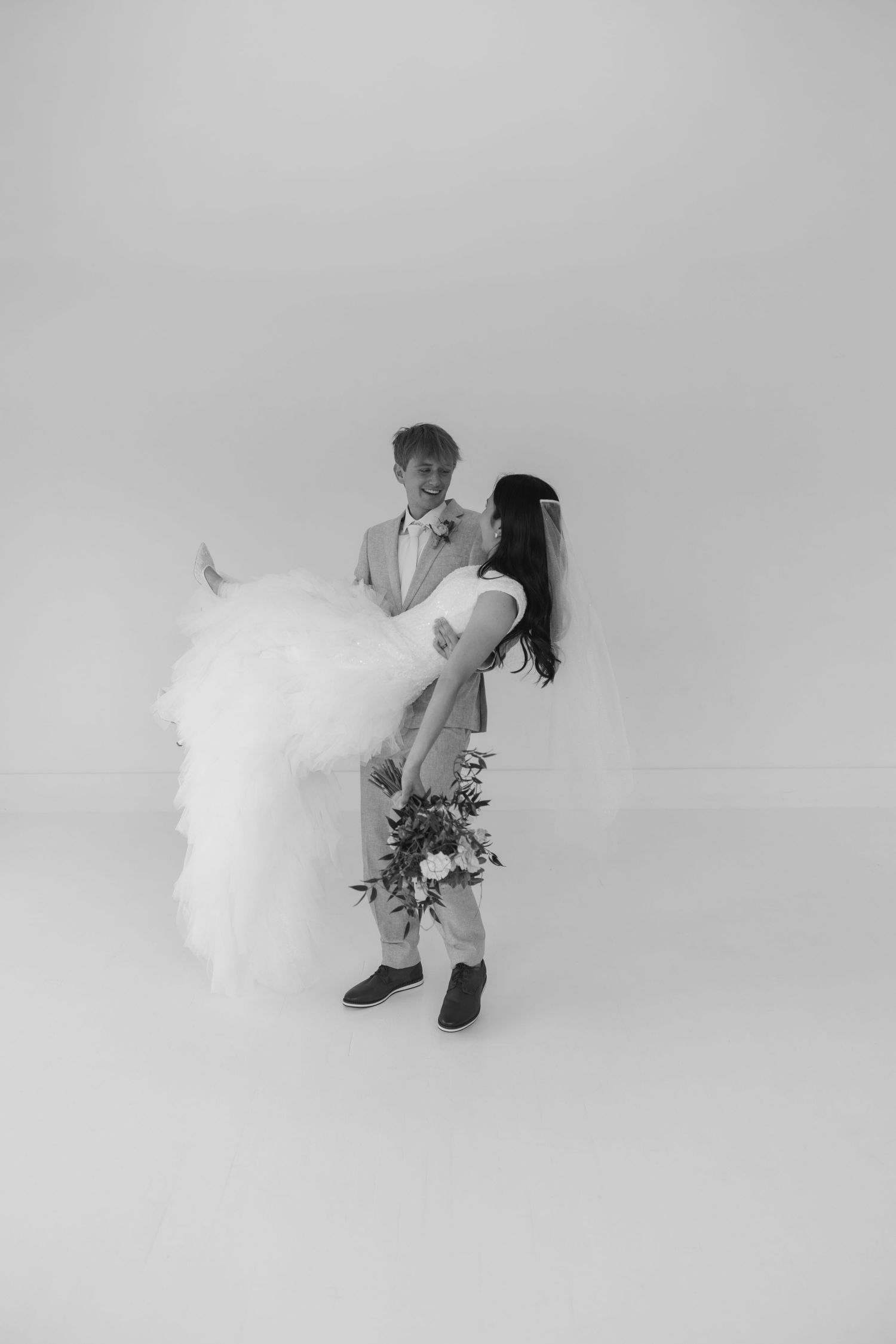 Black and white photo of groom lifting bride in wedding dress while she holds bouquet, studio backdrop.
