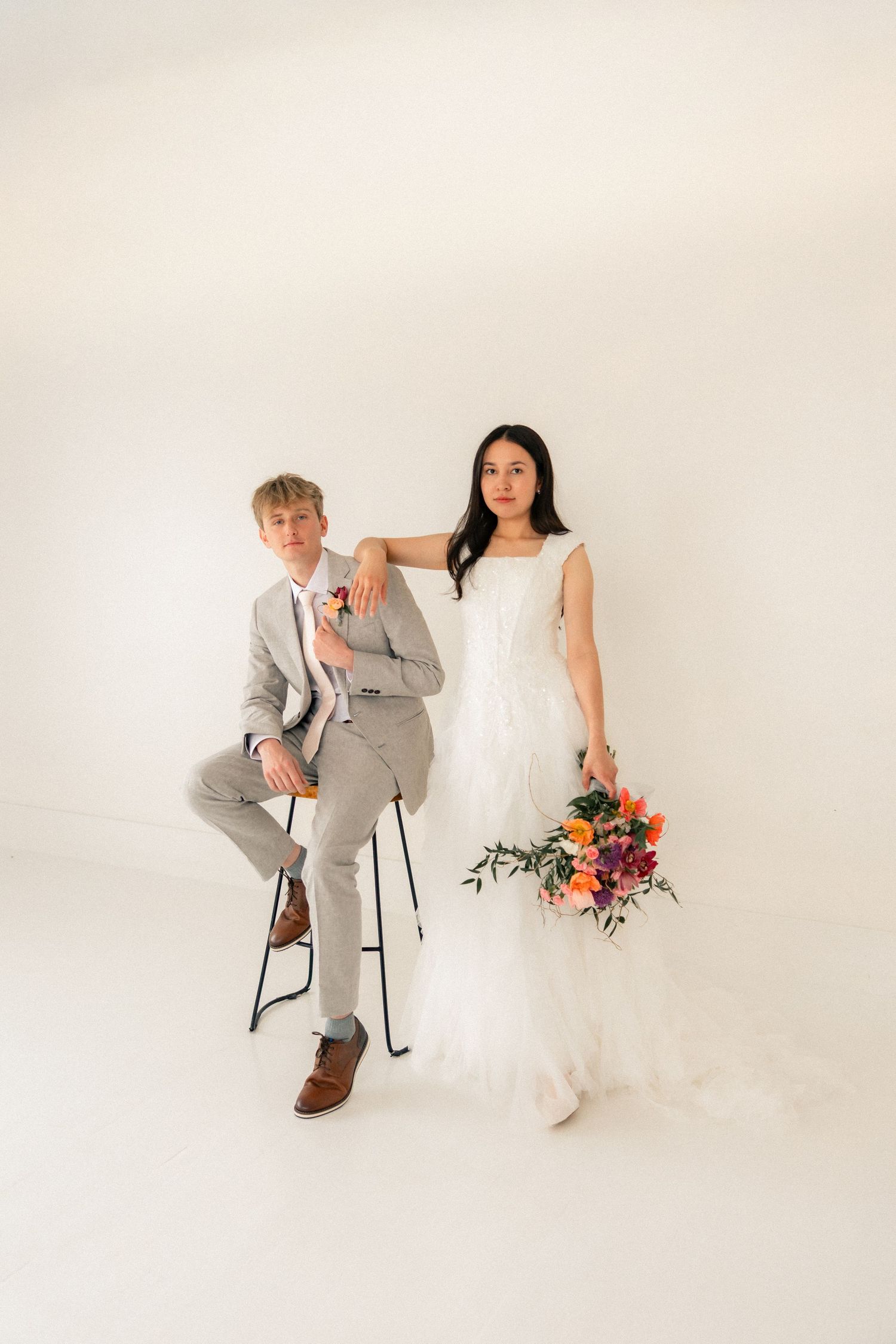 A bride in a white gown holding a colorful bouquet stands beside a groom in a gray suit seated on a stool.