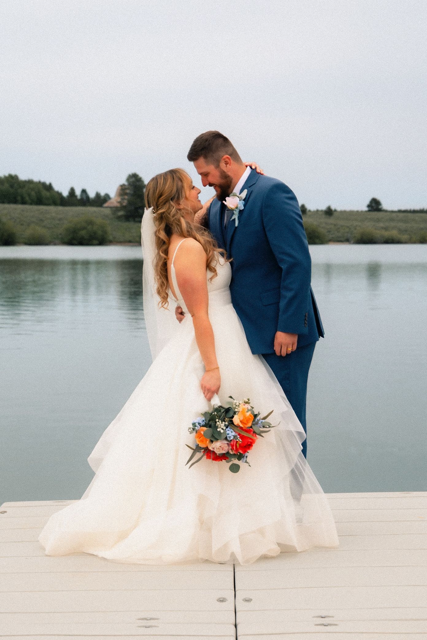 Bride and groom share a romantic moment on a dock by a lake, she in a white gown holding a colorful bouquet, he in a navy suit.