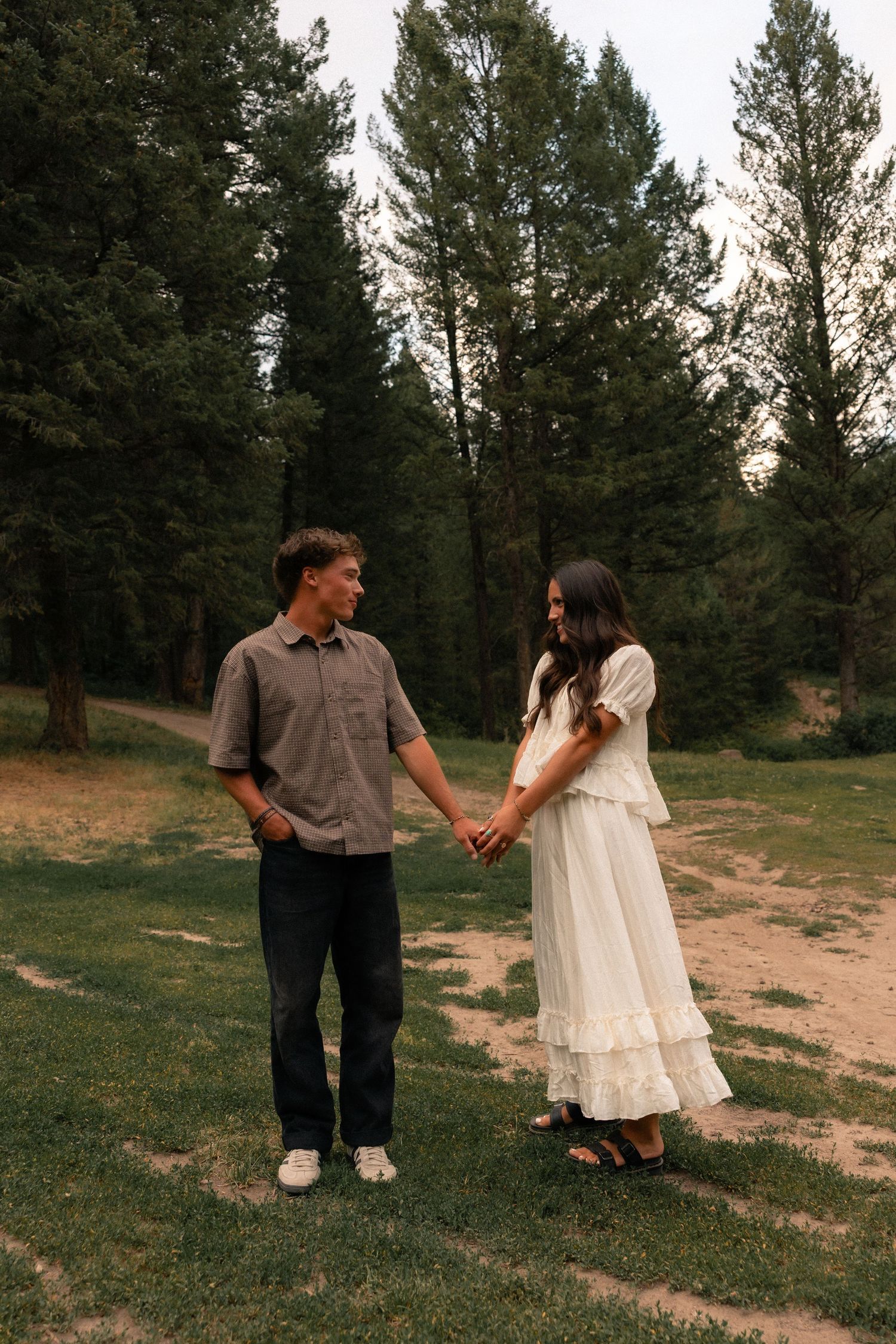 A couple holds hands in a forest clearing, the woman in a white dress and the man in a gray shirt and black pants.