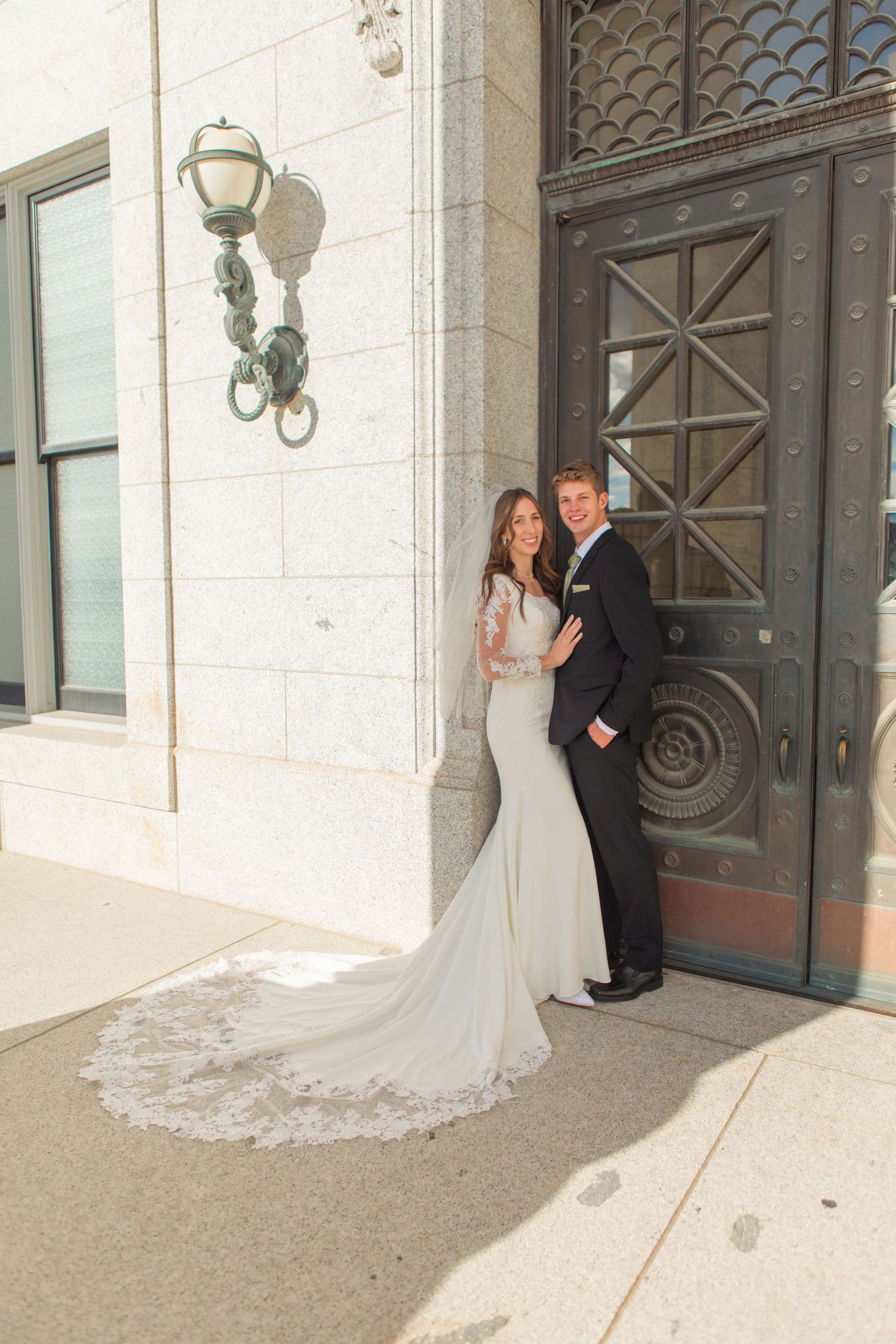 A couple in wedding attire poses in front of ornate metal doors at a grand entrance.