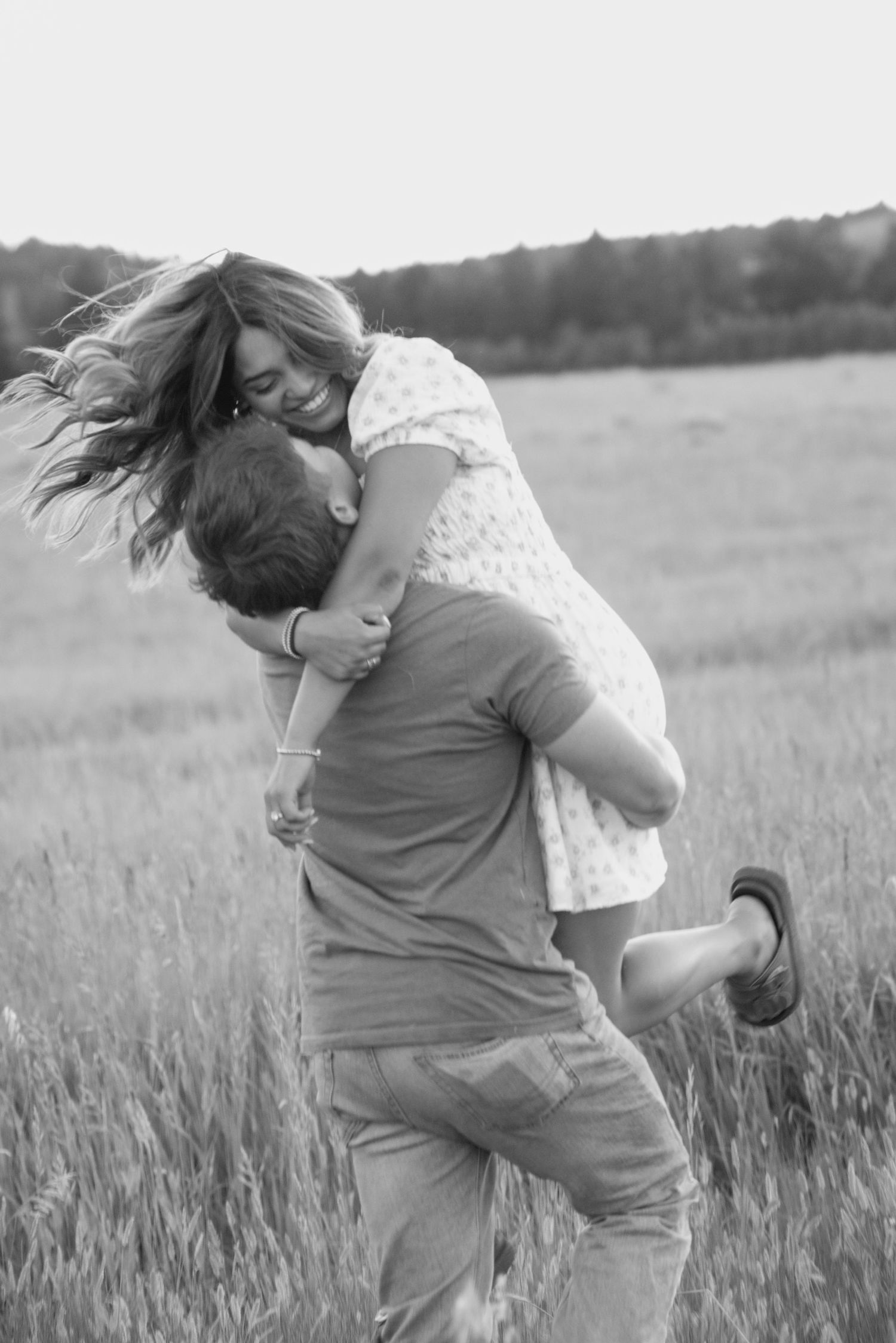 Black and white photo of playful couple dancing in rustic field at dusk.
