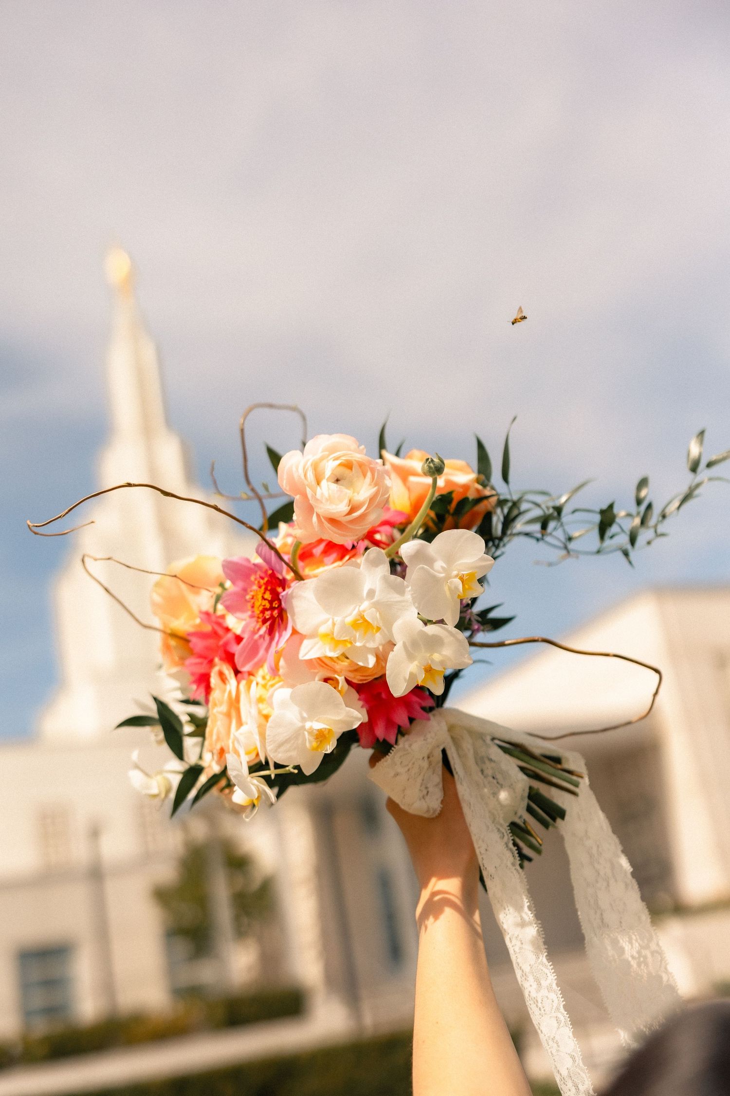 A bridal bouquet of pink, peach, and white flowers held up against a blue sky with a white temple in the background.