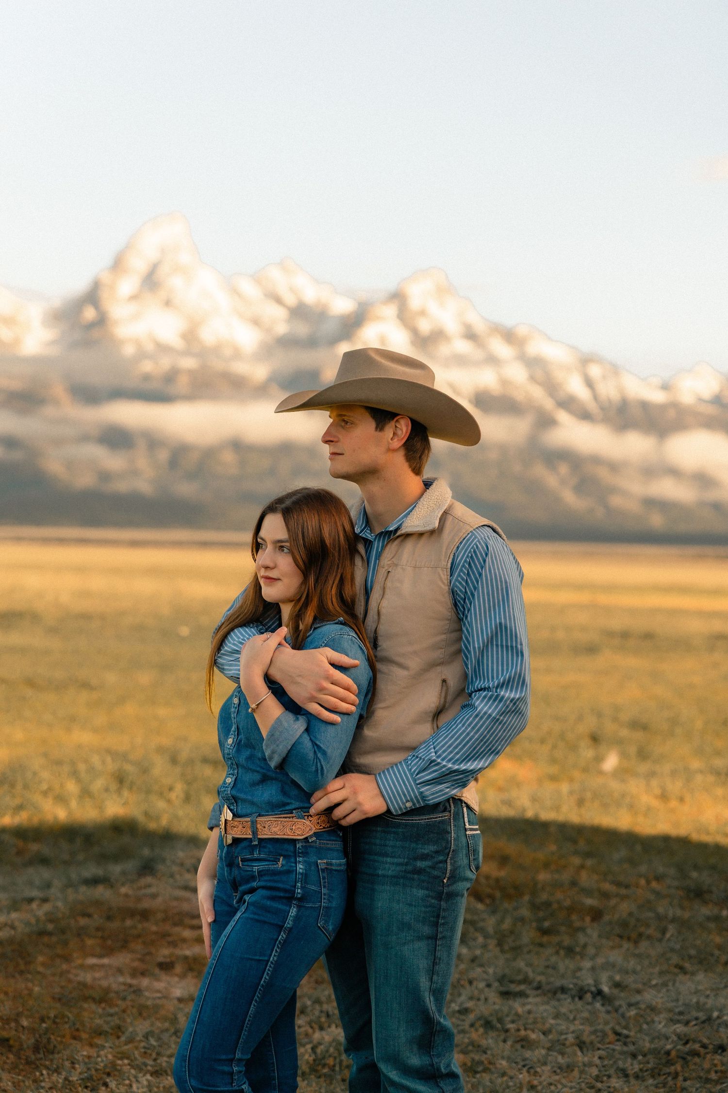 A couple embraces in a golden field with snow-capped Teton mountains behind them, man wearing cowboy hat and vest.