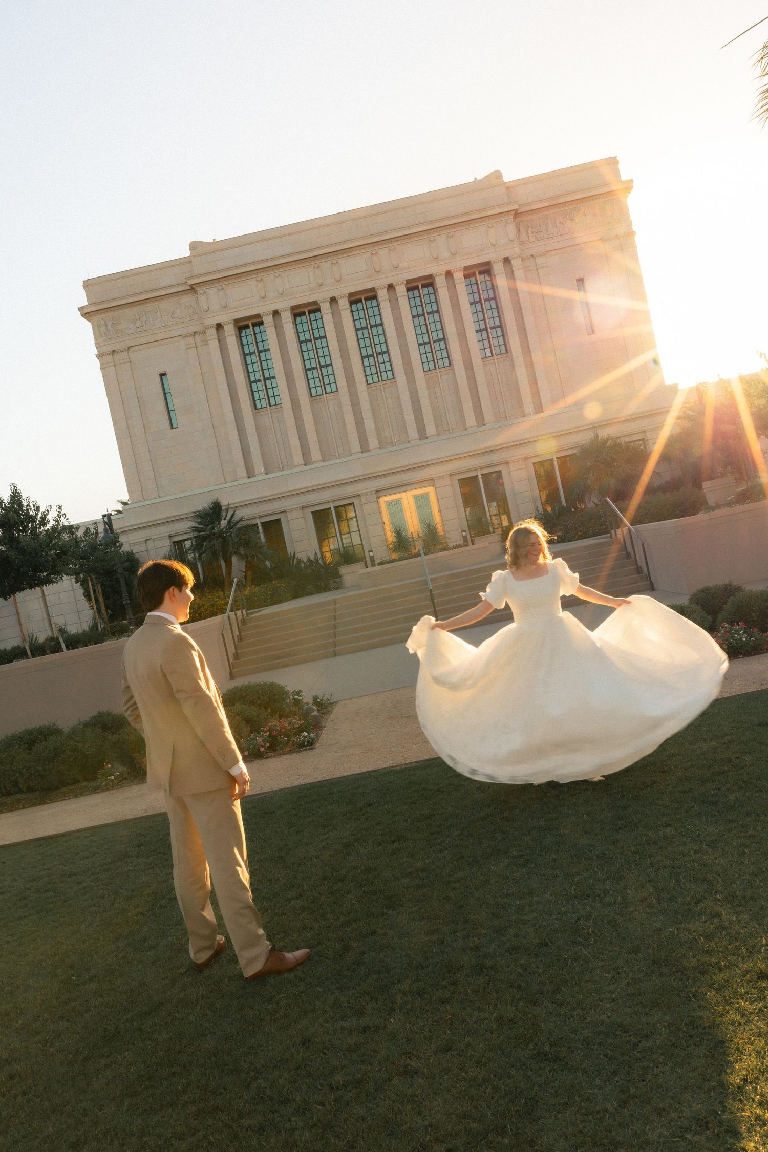 Bride in white gown twirling before groom at sunset outside a grand temple on their wedding day.