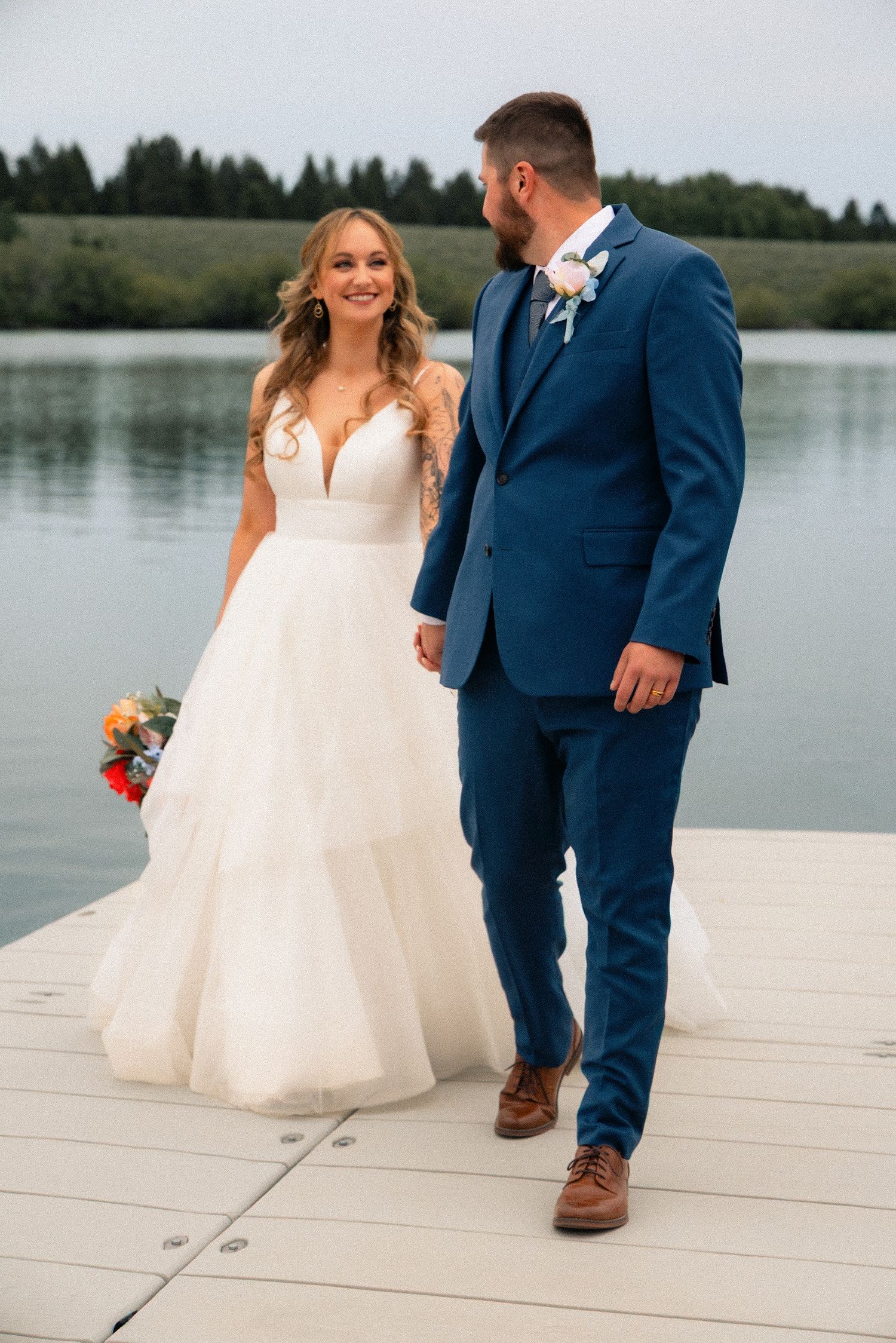 Bride in white gown and groom in blue suit walk hand in hand on a dock by a serene lake.