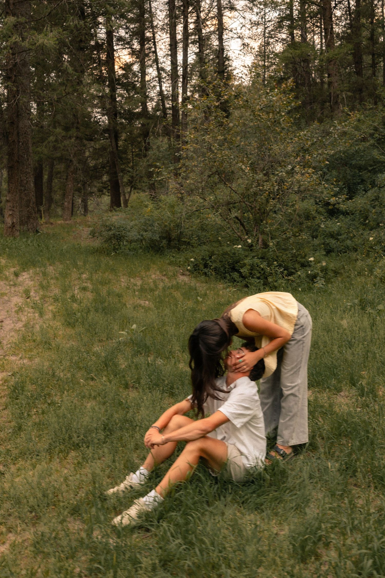 Two women share a tender moment on grass in a forest clearing, one sitting while the other leans down to embrace her.
