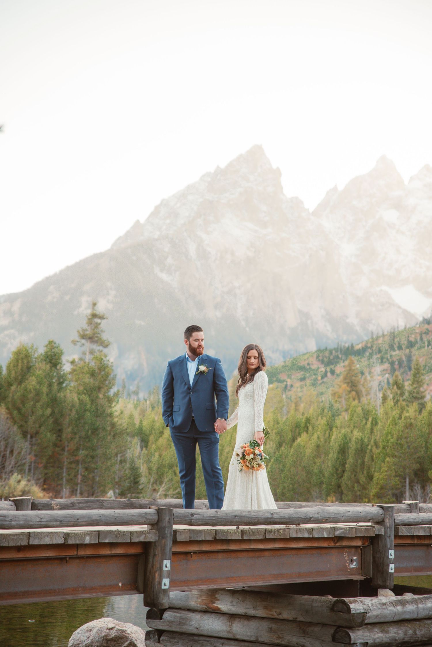 Couple in wedding attire standing on wooden dock with majestic mountain backdrop in golden light.