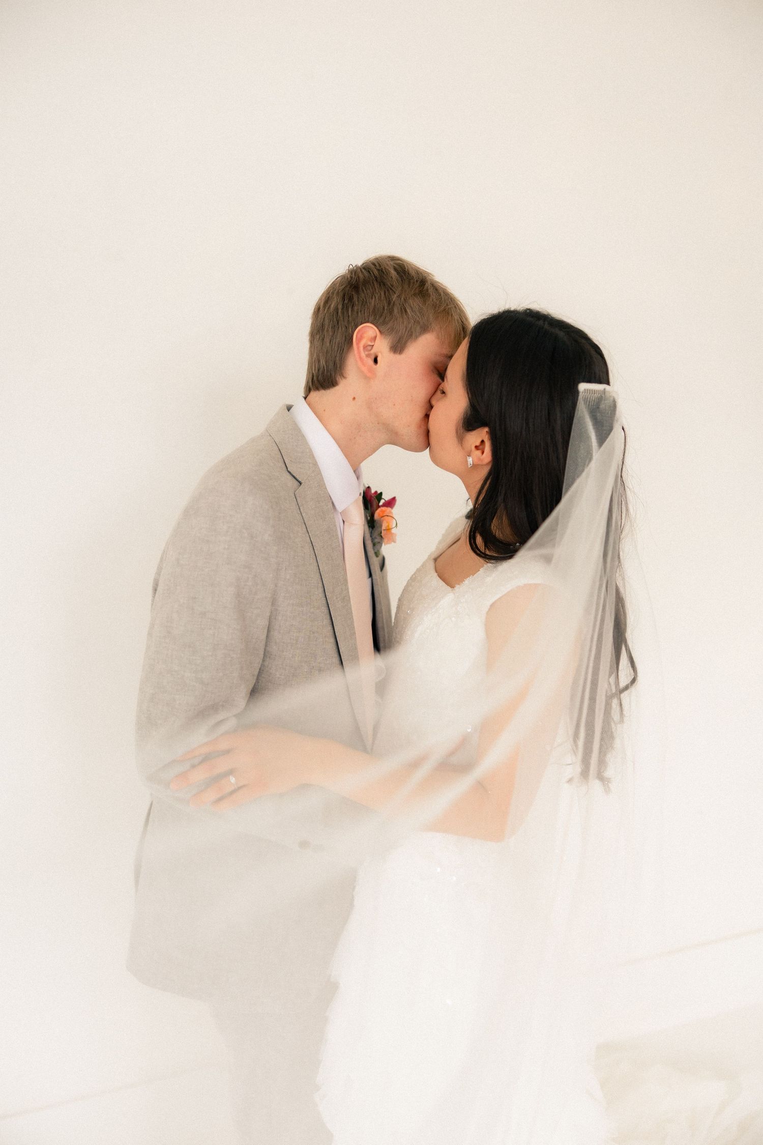 A bride and groom share a romantic kiss, with her white veil draped around them, as he wears a light gray suit.