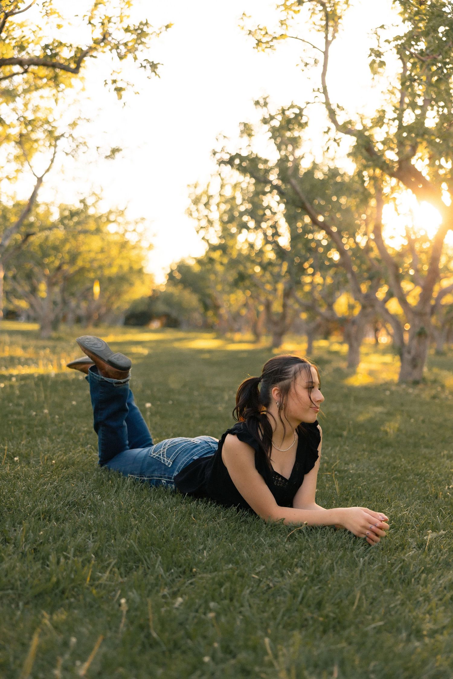 Young woman lying on green grass in a sunny park at golden hour, wearing jeans and a black top.