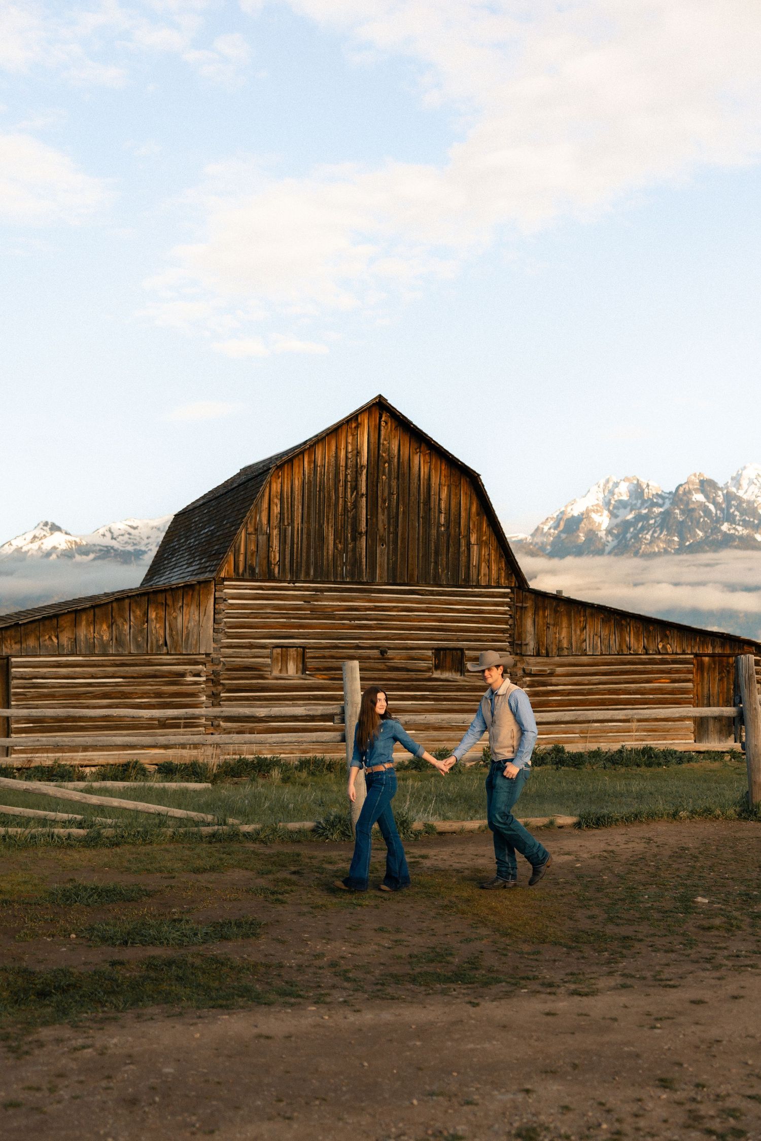 Couple holding hands walking in front of a rustic wooden barn with snow-capped Grand Teton mountains in the background.
