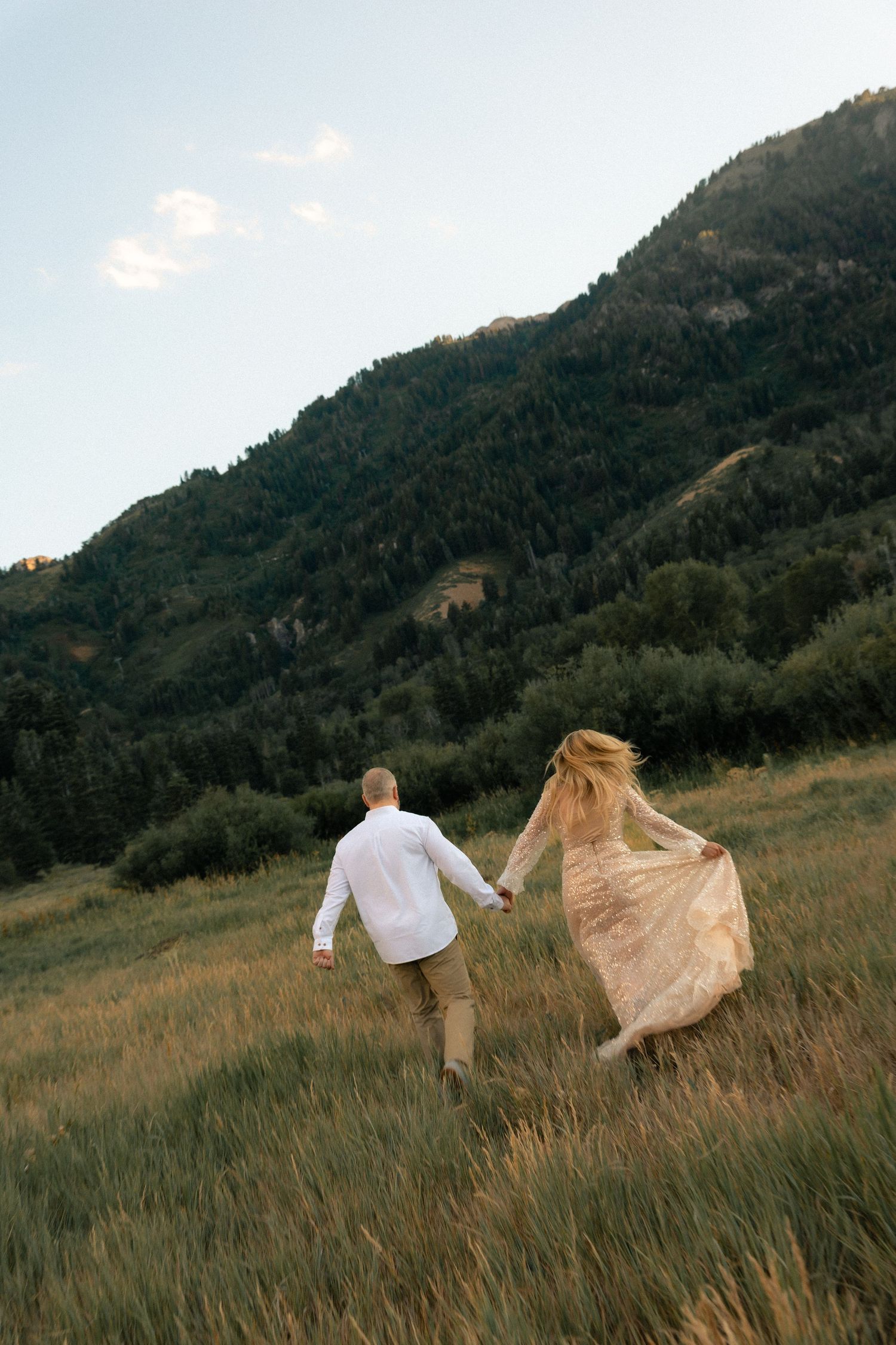 Couple holding hands running through a grassy mountain meadow, woman in flowing dress, man in white shirt, lush green hills.