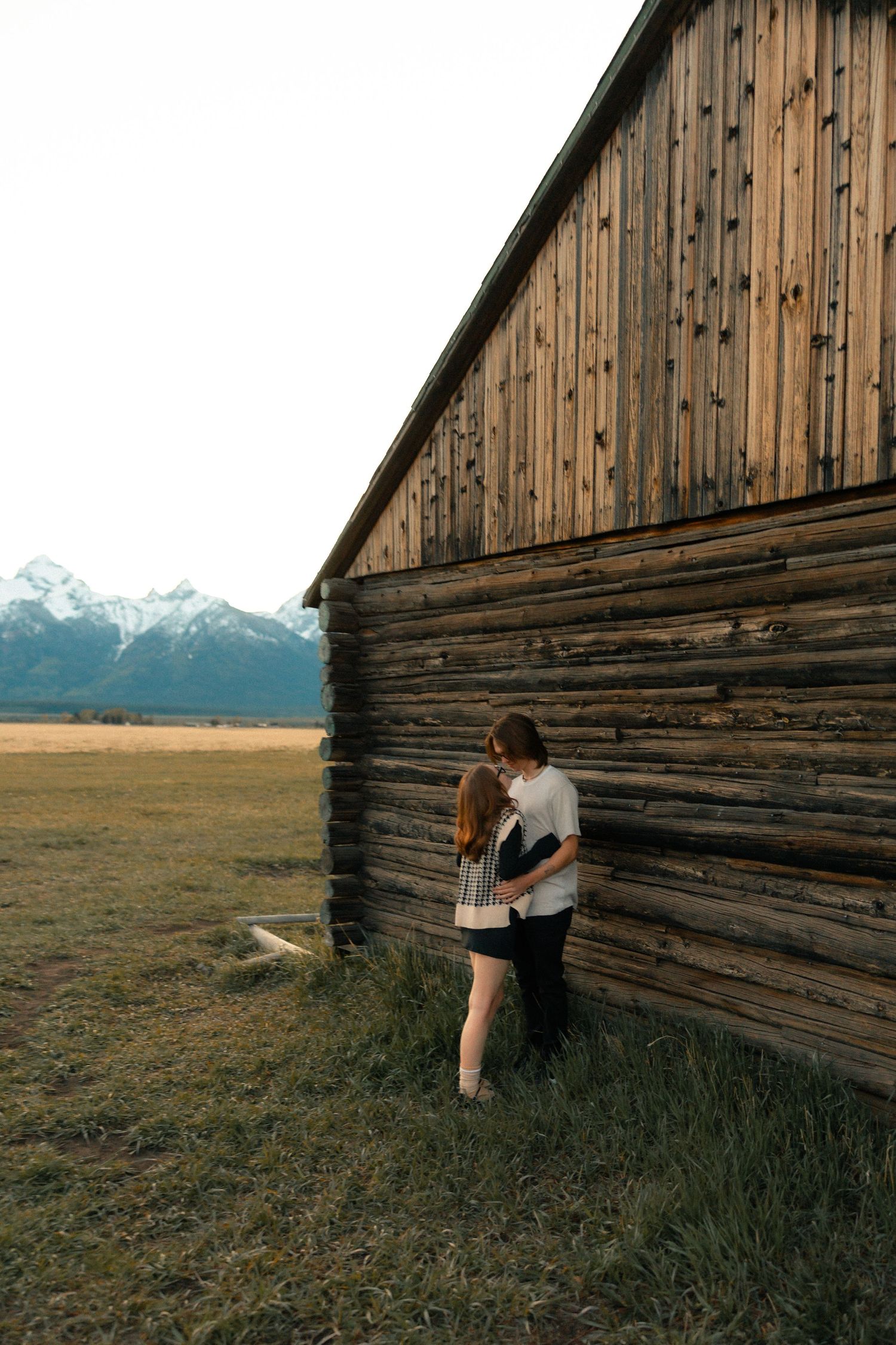 Couple embracing against rustic wooden barn with snow-capped mountains in background on grassy field.