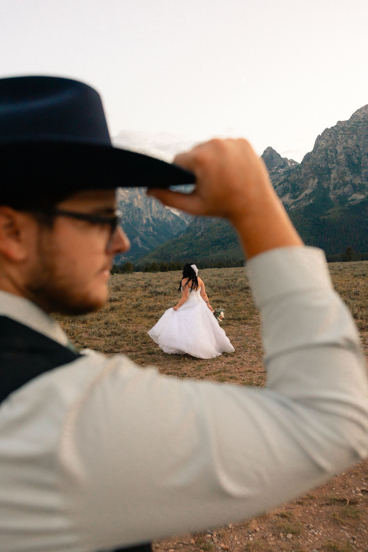 Groom in cowboy hat and glasses tips hat as bride in white gown walks through mountain meadow at sunset.
