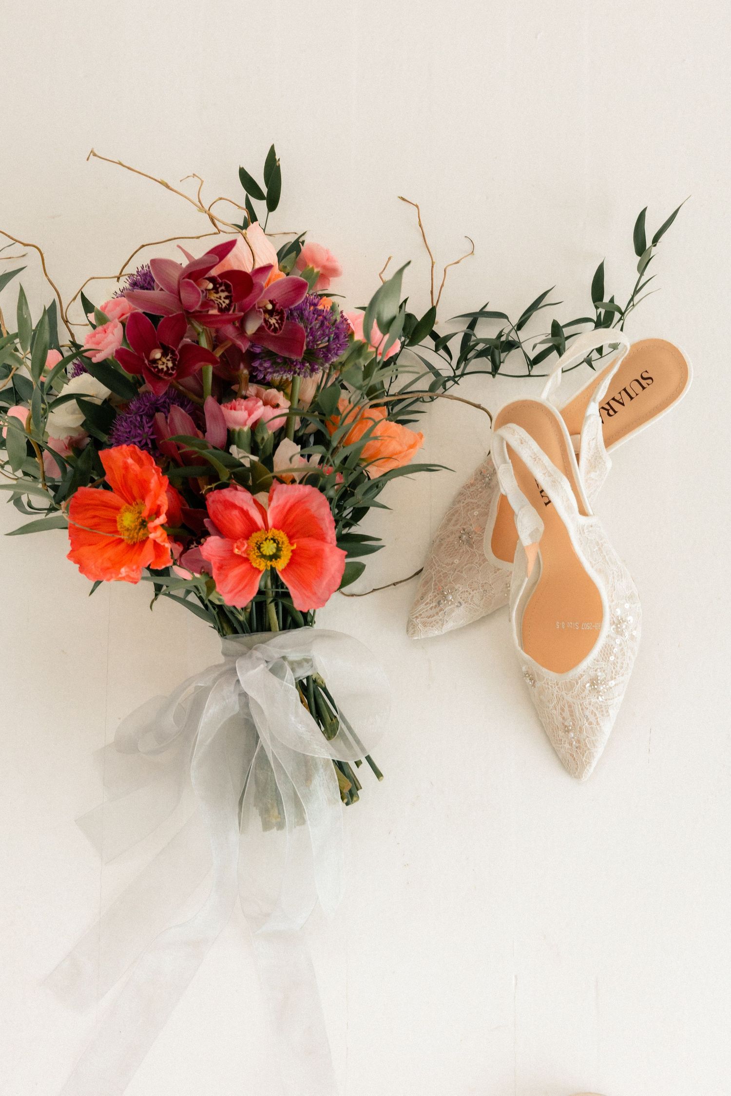 Bridal bouquet with orange poppies and purple flowers beside lace slingback wedding heels on white background.
