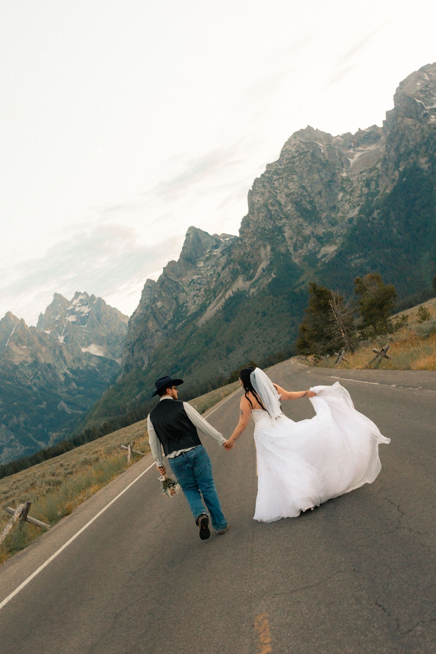Bride and groom walk hand-in-hand down a mountain road with Grand Teton peaks in the background.