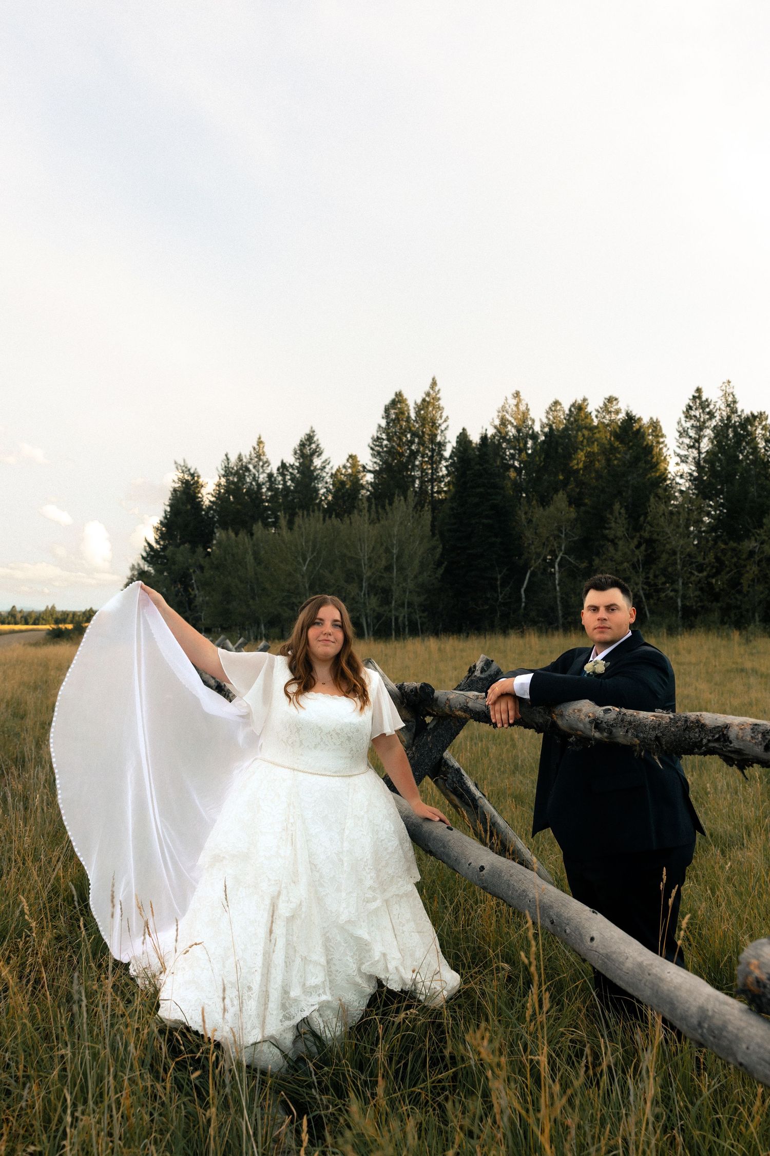 Bride in white lace gown holds veil as groom in black suit leans on rustic fence in meadow with pine trees.