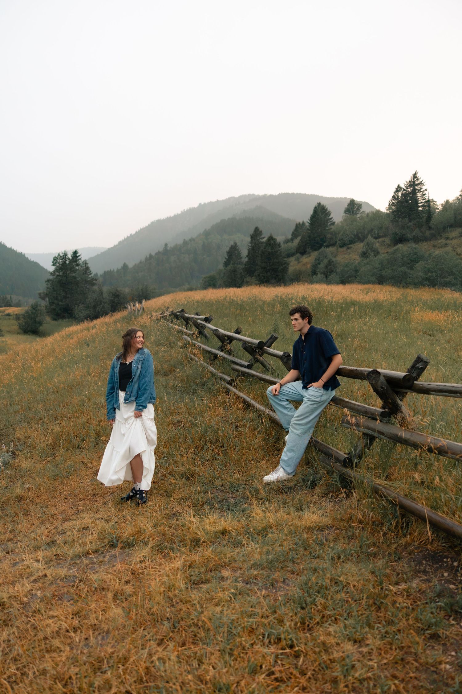 Couple poses by rustic wooden fence in mountain meadow, woman in white skirt and denim jacket, man in blue shirt.