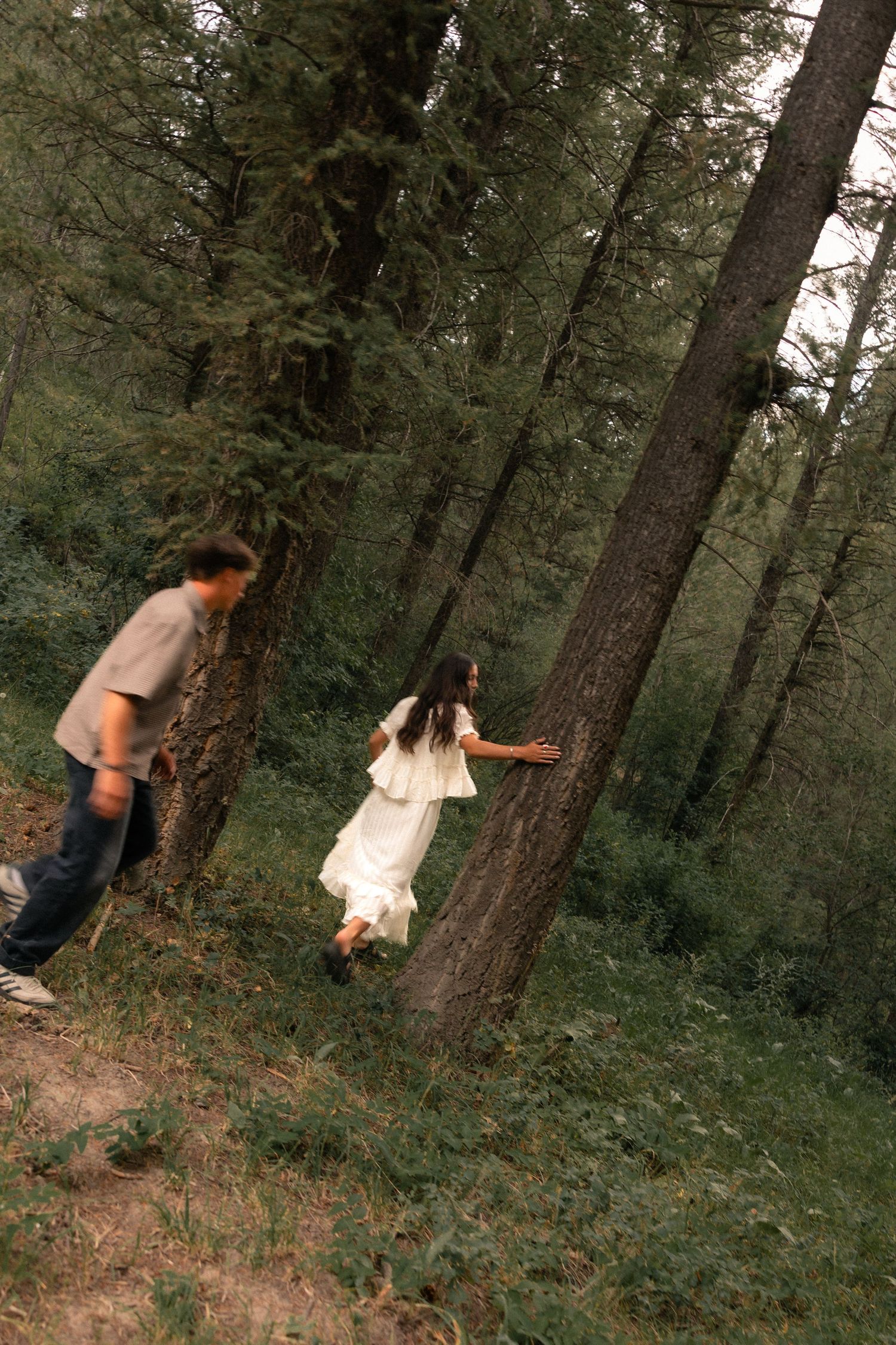 A couple plays in a lush green forest, woman in white dress touching a tree trunk while man approaches from behind.