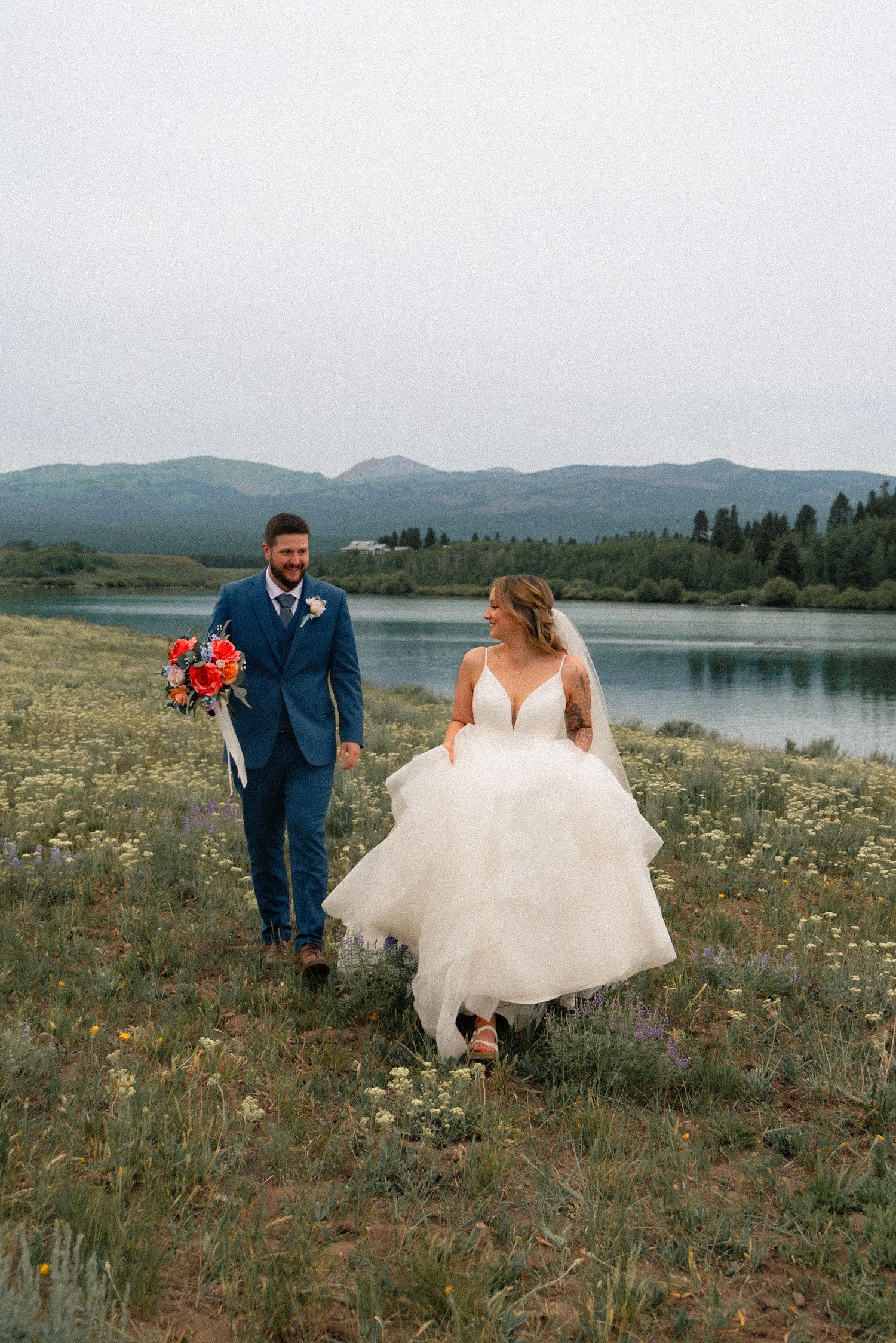 Bride and groom walk hand-in-hand through a wildflower meadow beside a mountain lake, bride in white gown, groom in blue suit.
