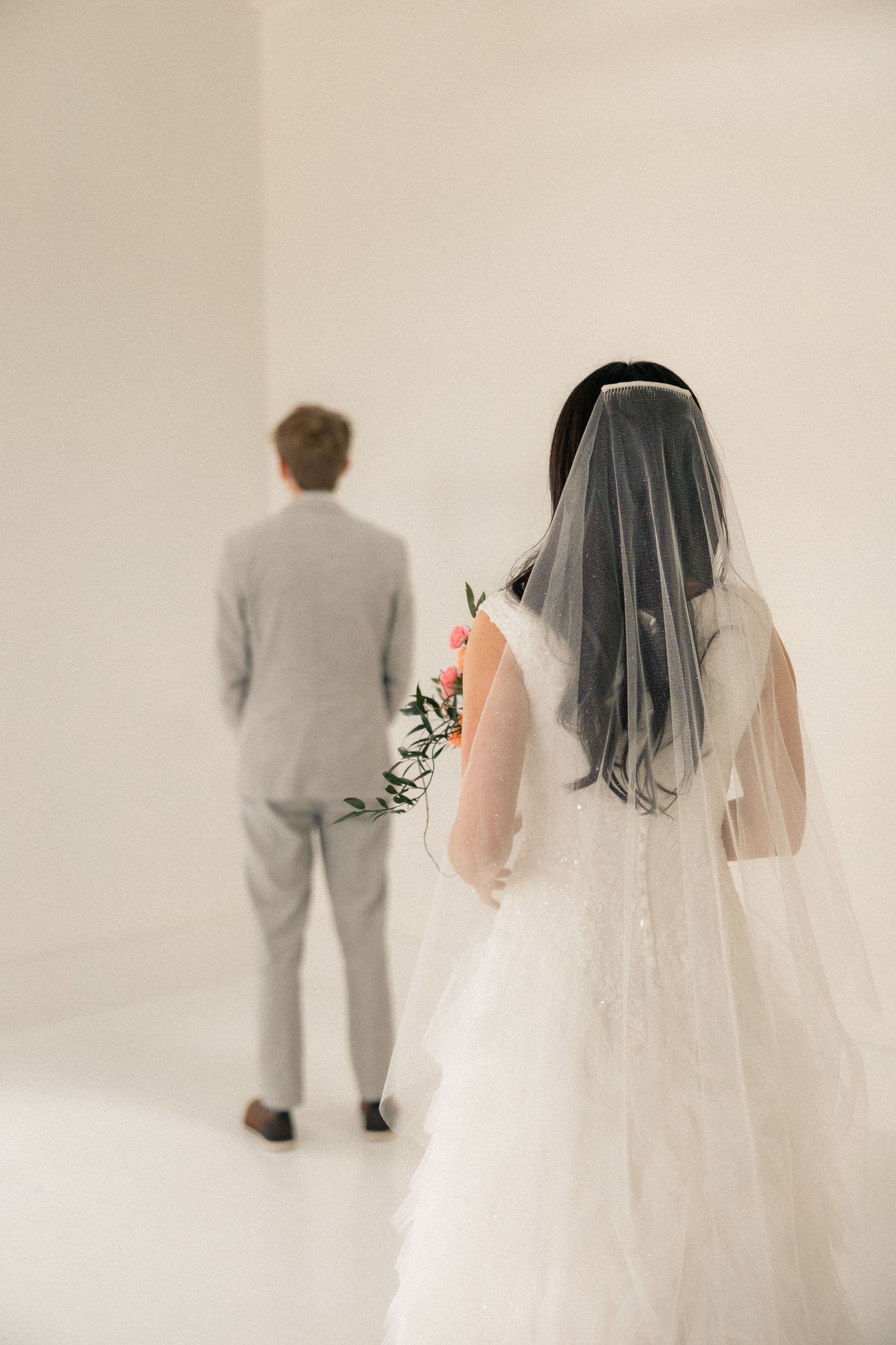 Bride in white lace gown and veil holding pink flowers approaches groom in gray suit during wedding first look.