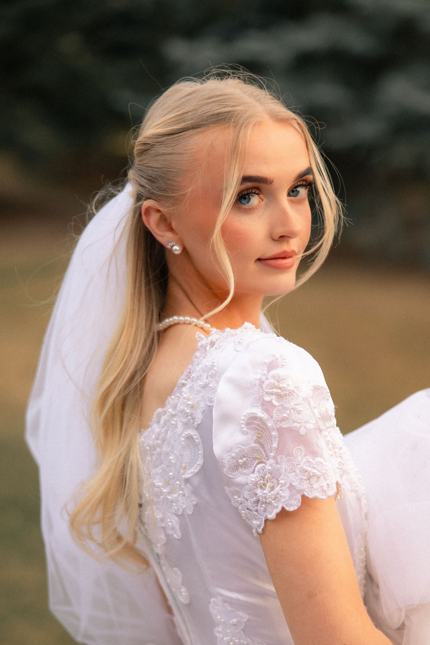 Young blonde bride in white lace wedding dress and veil, looking over shoulder outdoors.