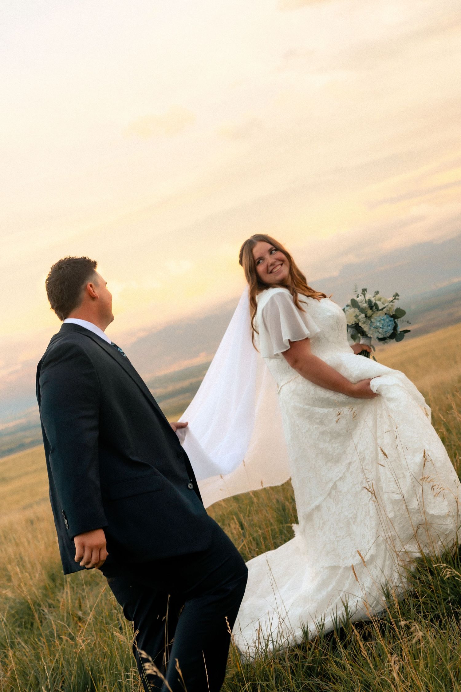 A bride in a white lace gown holds her bouquet while smiling at her groom in a black suit during a golden sunset.