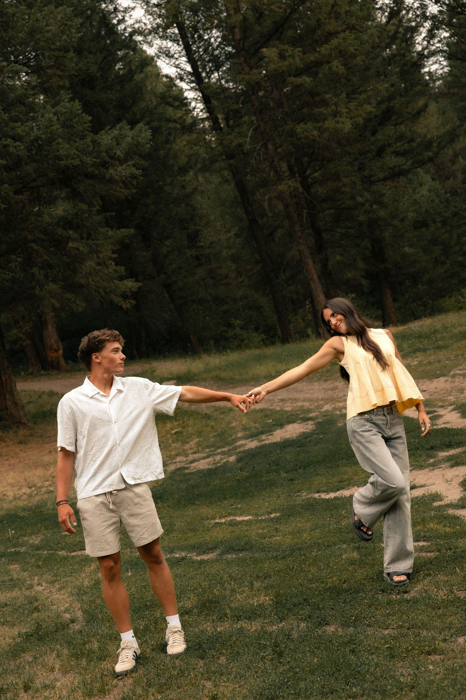 Couple holding hands in a lush green park, man in white shirt and shorts, woman in yellow top and gray jeans.