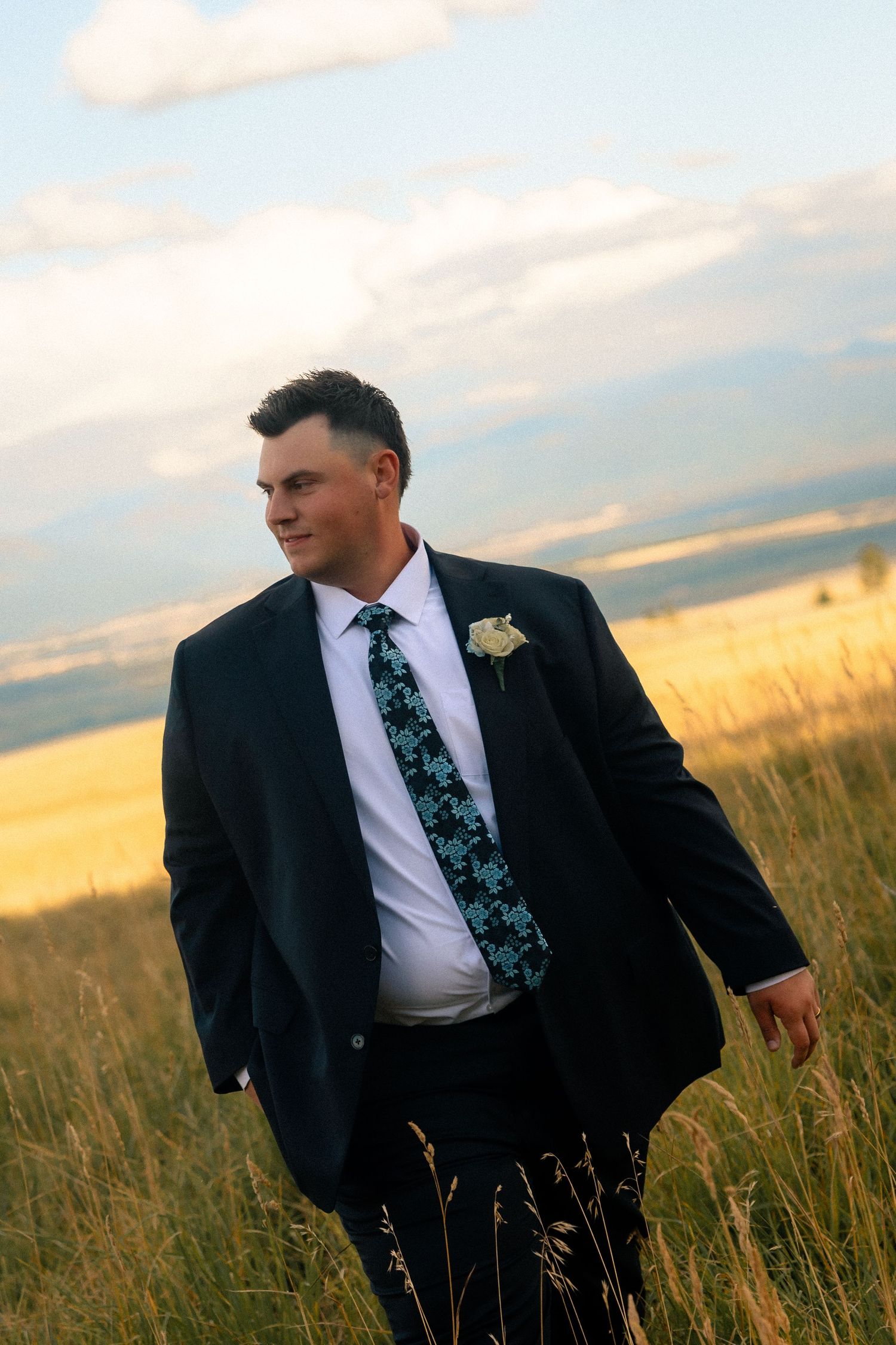 A groom in a navy suit and floral tie with boutonniere stands in a golden field under a cloudy sky.