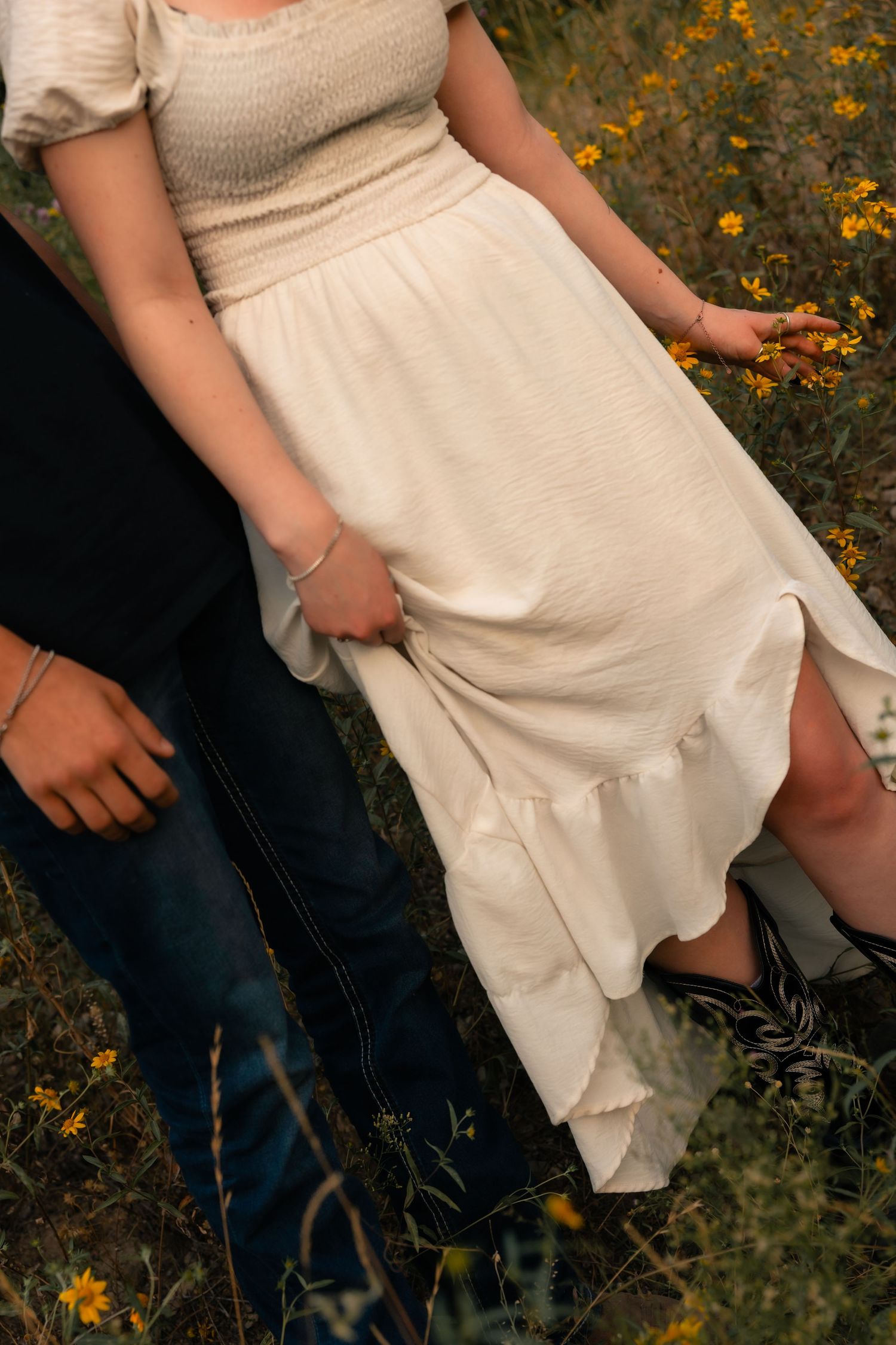 Couple standing in a wildflower field, woman wearing a white smocked midi dress with yellow flowers in background.
