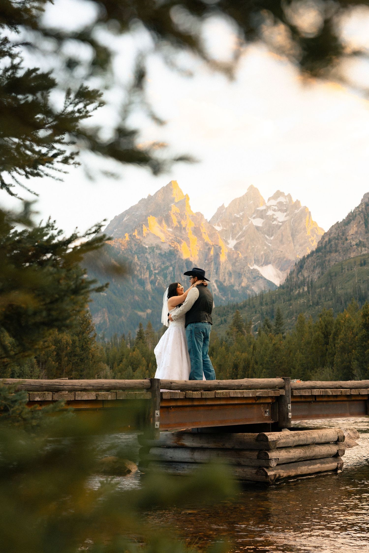 Couple embracing on a wooden bridge with Grand Teton mountains glowing at sunset behind them.