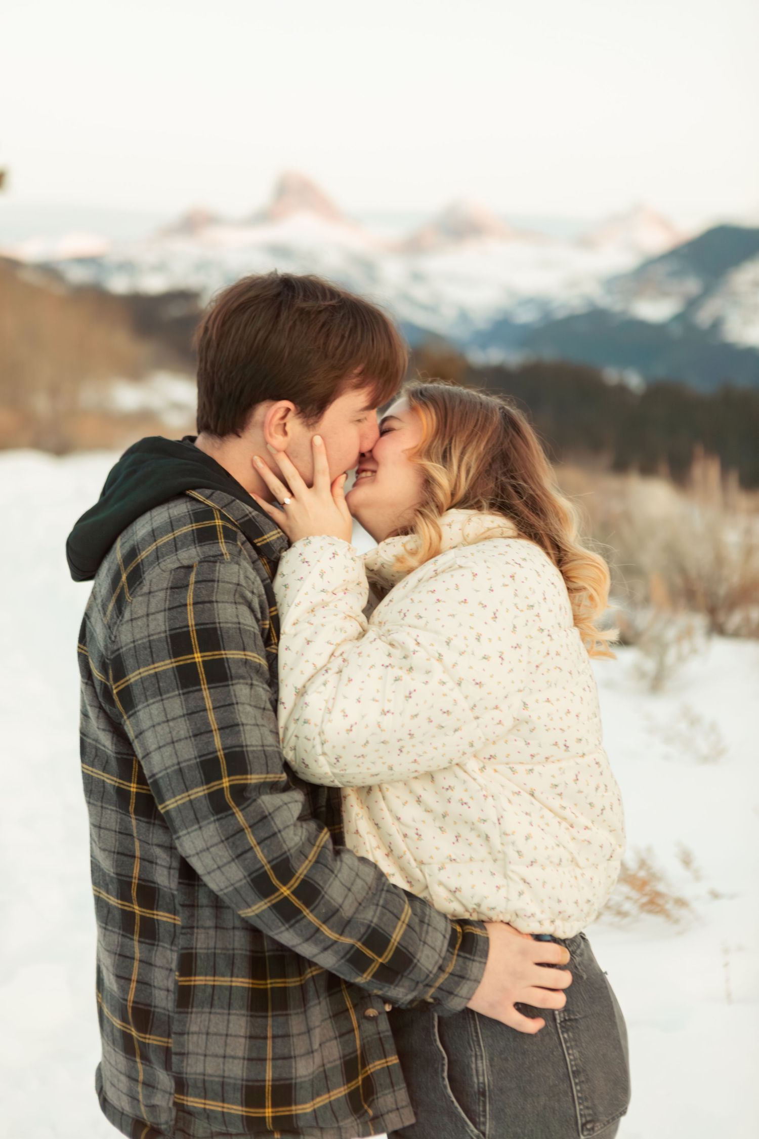 A romantic winter engagement photoshoot featuring a couple embracing in the snow with mountains in the background.