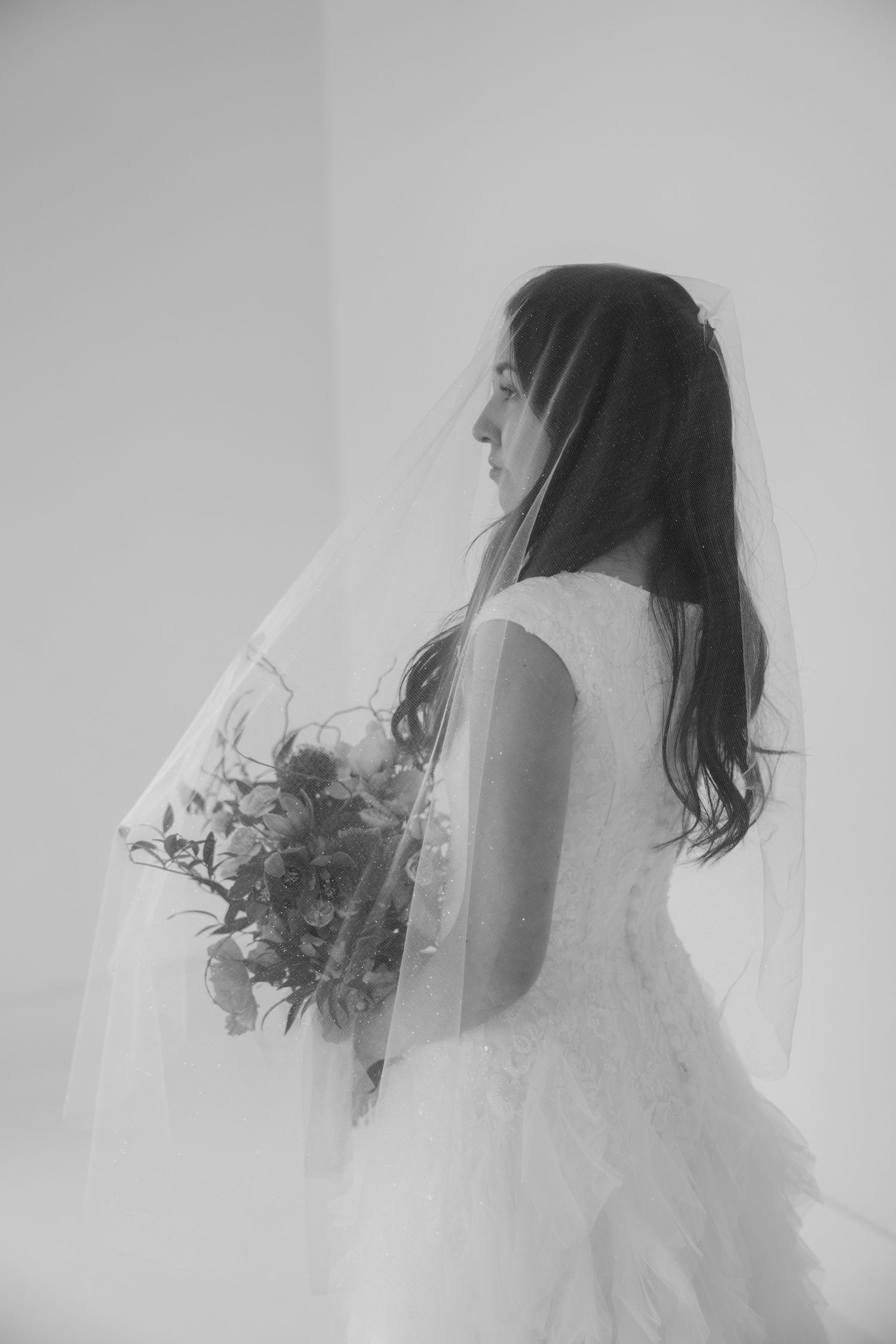Black and white photo of a bride in a wedding gown and veil holding a bouquet, viewed from behind.
