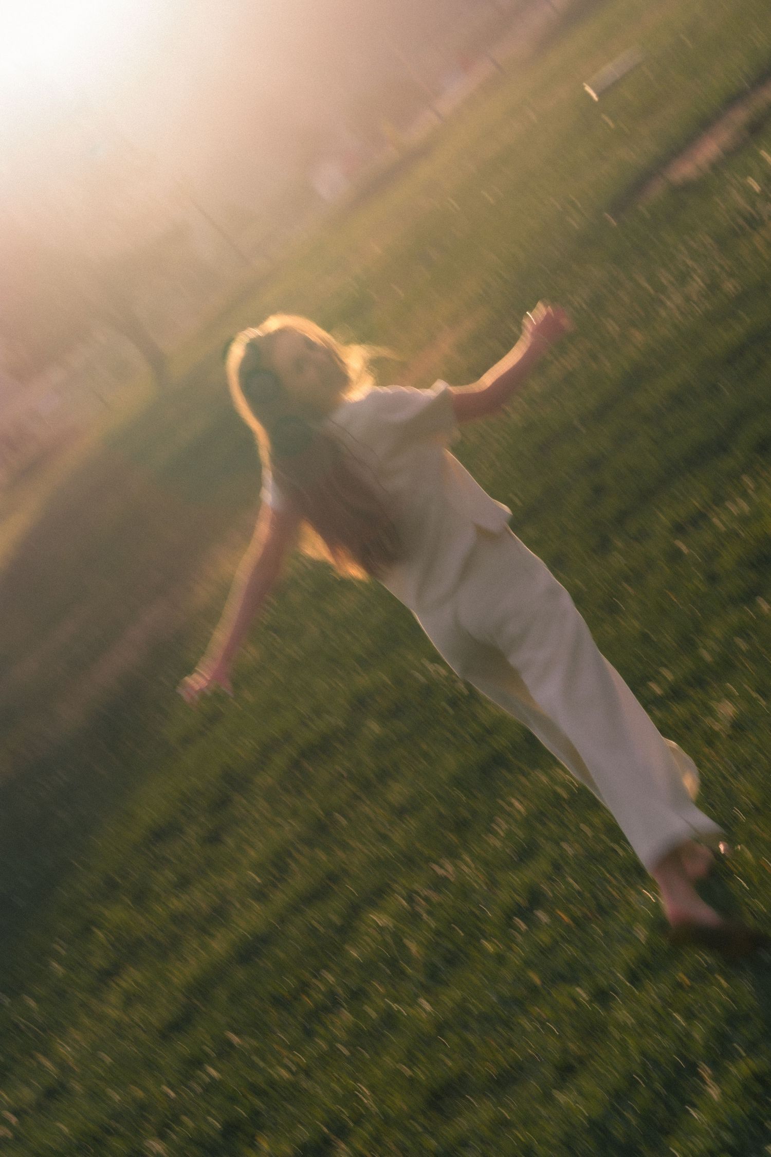 Blurred motion shot of a woman in white outfit spinning in a sunlit green field with golden lens flare.