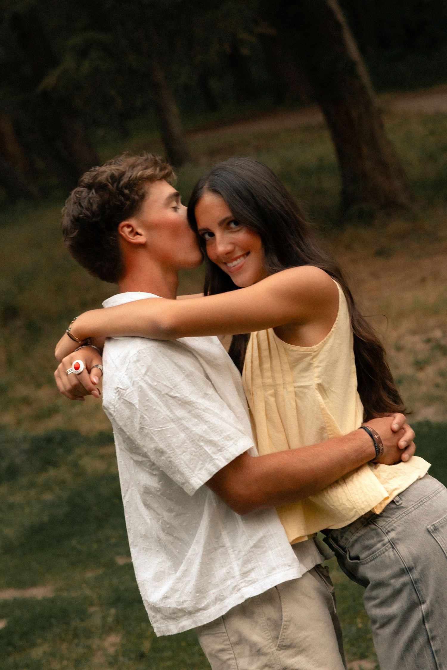 Young couple embracing outdoors, woman smiling as man kisses her cheek in a lush green park setting.